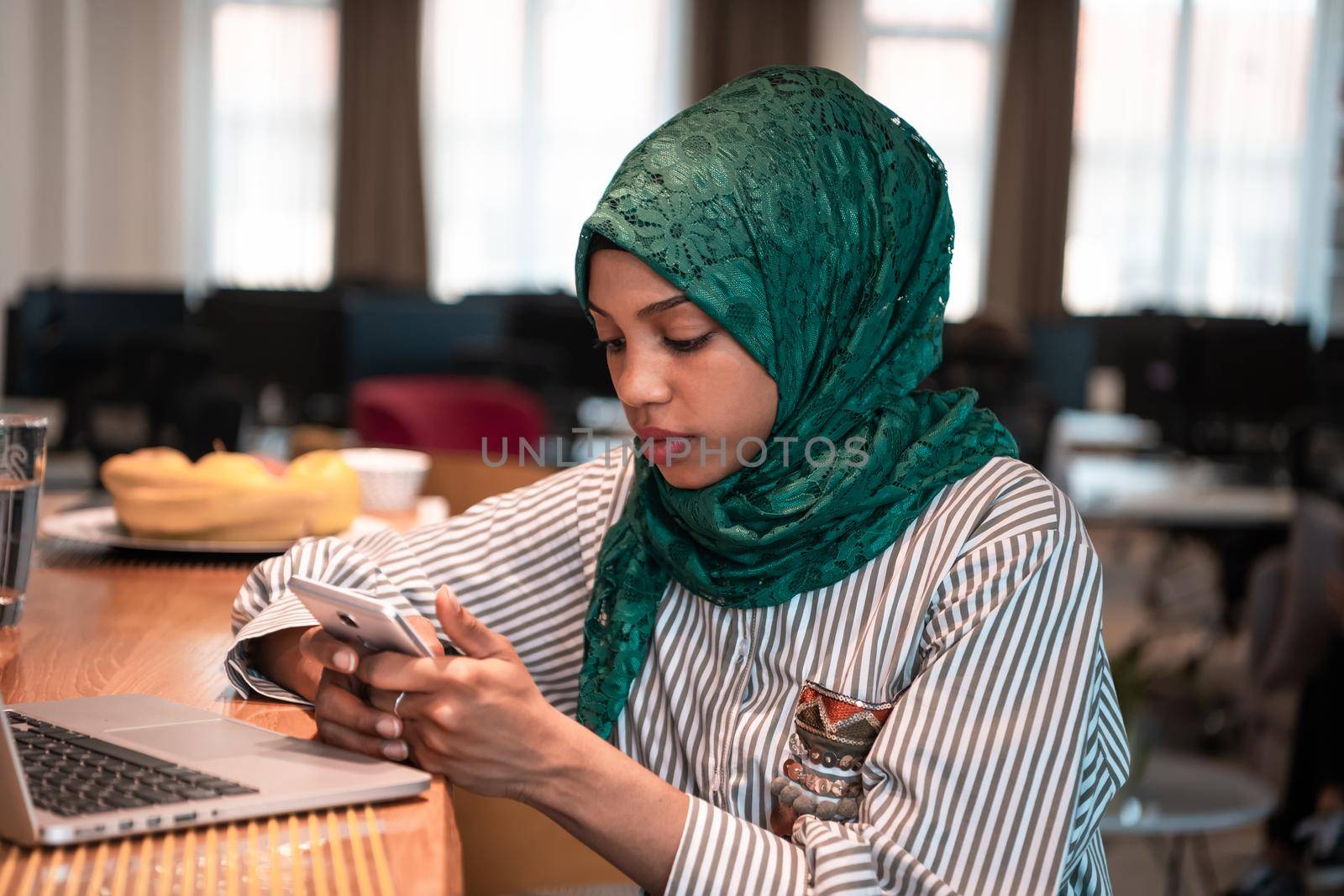 African Muslim business woman wearing a green hijab using smartphone while working on laptop computer in relaxation area at modern open plan startup office. by dotshock