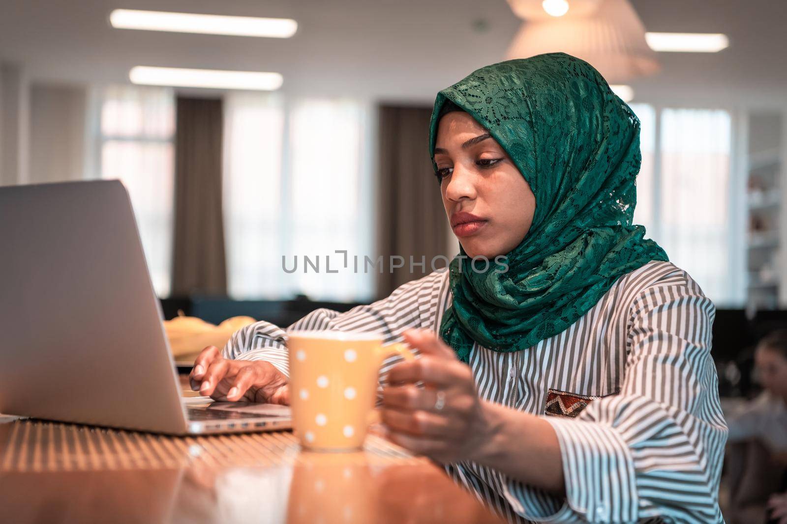 African Muslim businesswoman wearing a green hijab drinking tea while working on laptop computer in relaxation area at modern open plan startup office. High-quality photo