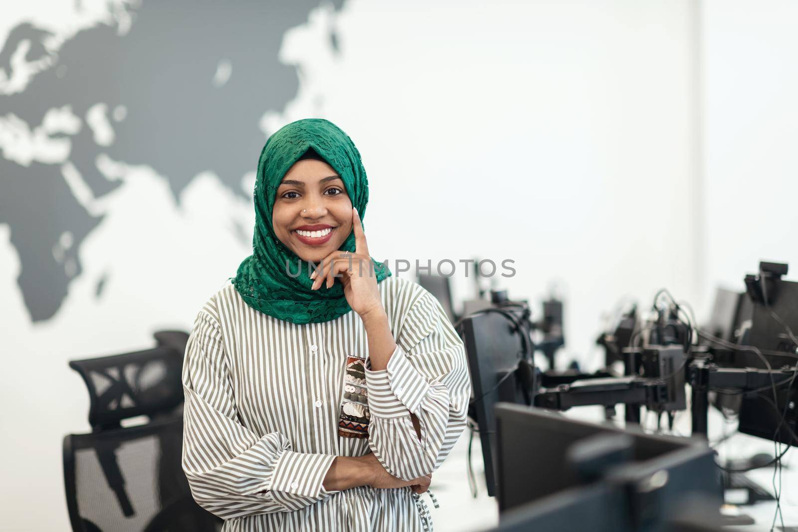 Portrait of Muslim black female software developer with green hijab standing at modern open plan startup office. Selective focus. High-quality photo