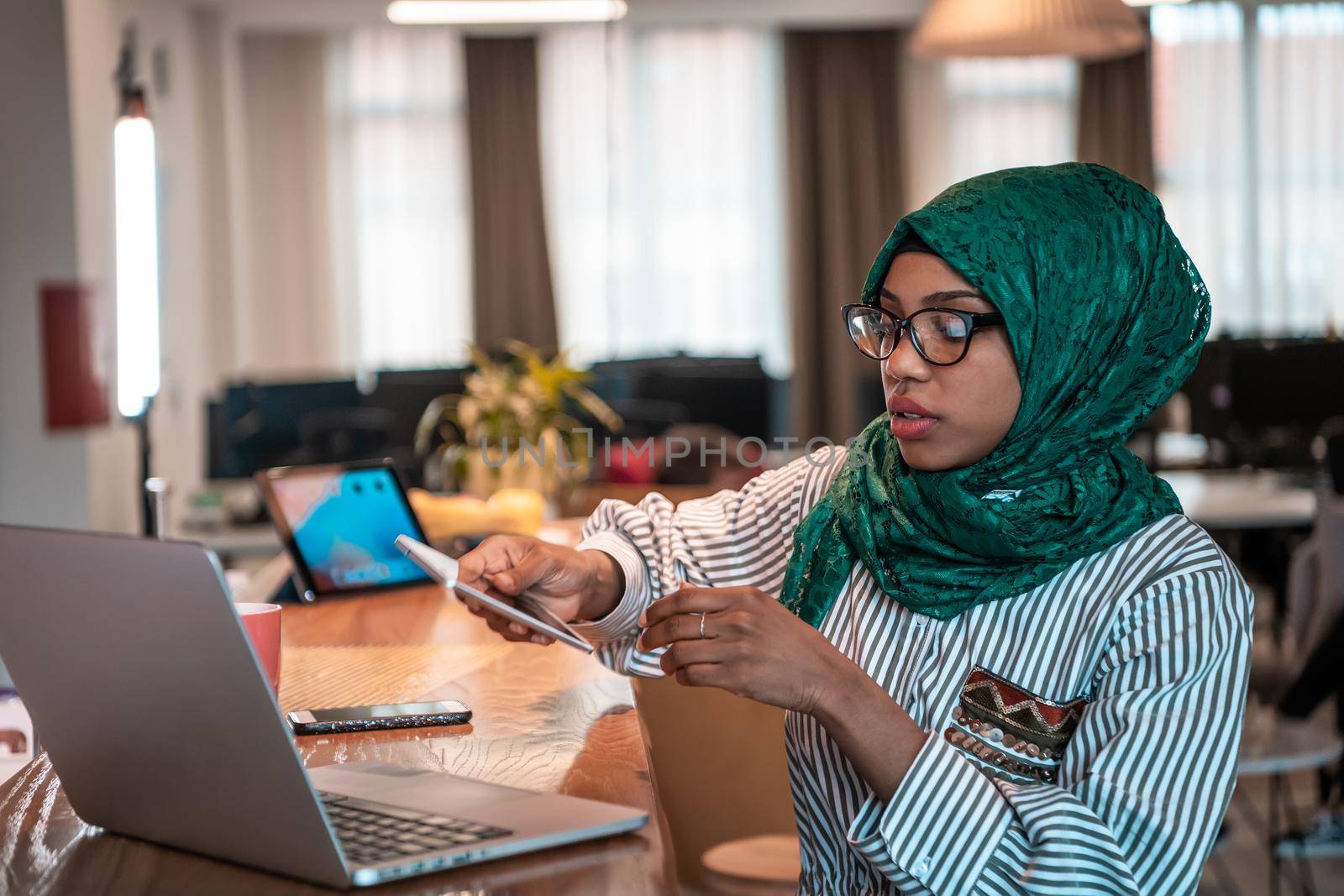 Smiling young woman talk on smartphone work on laptop look in distance thinking. Happy African American customer client using modern gadgets cellphone and computer at the home office. High-quality photo