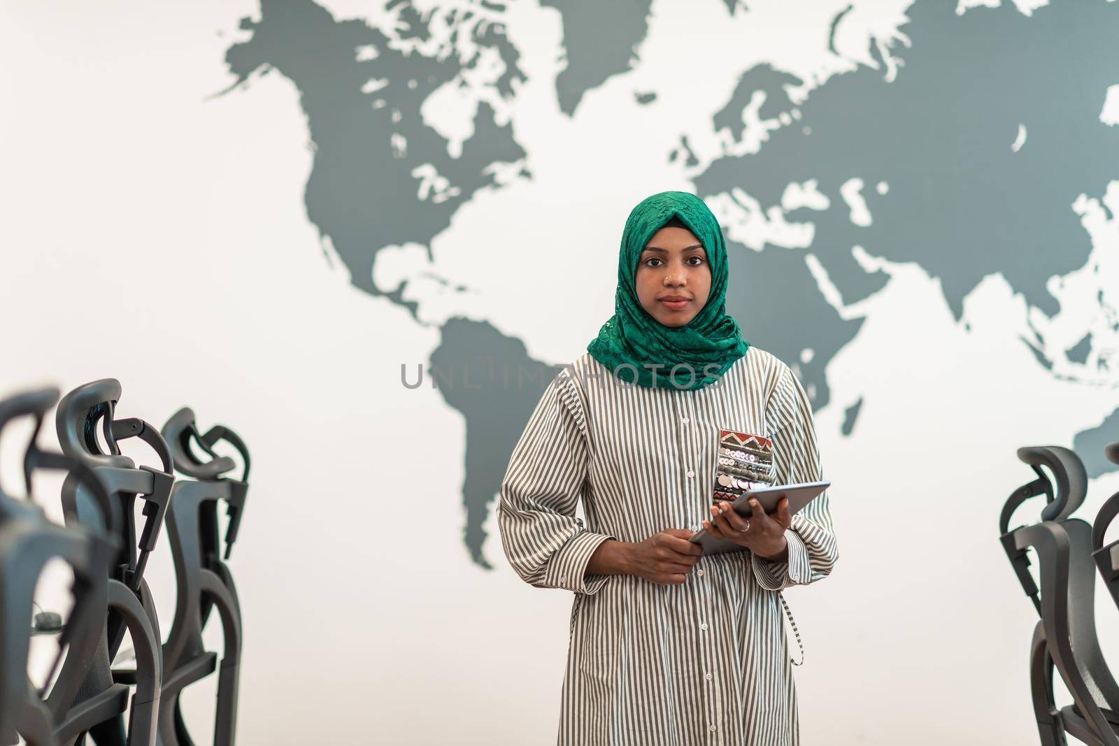 Portrait of muslim female software developer with green hijab holding tablet computer while standing at modern open plan startup office by dotshock