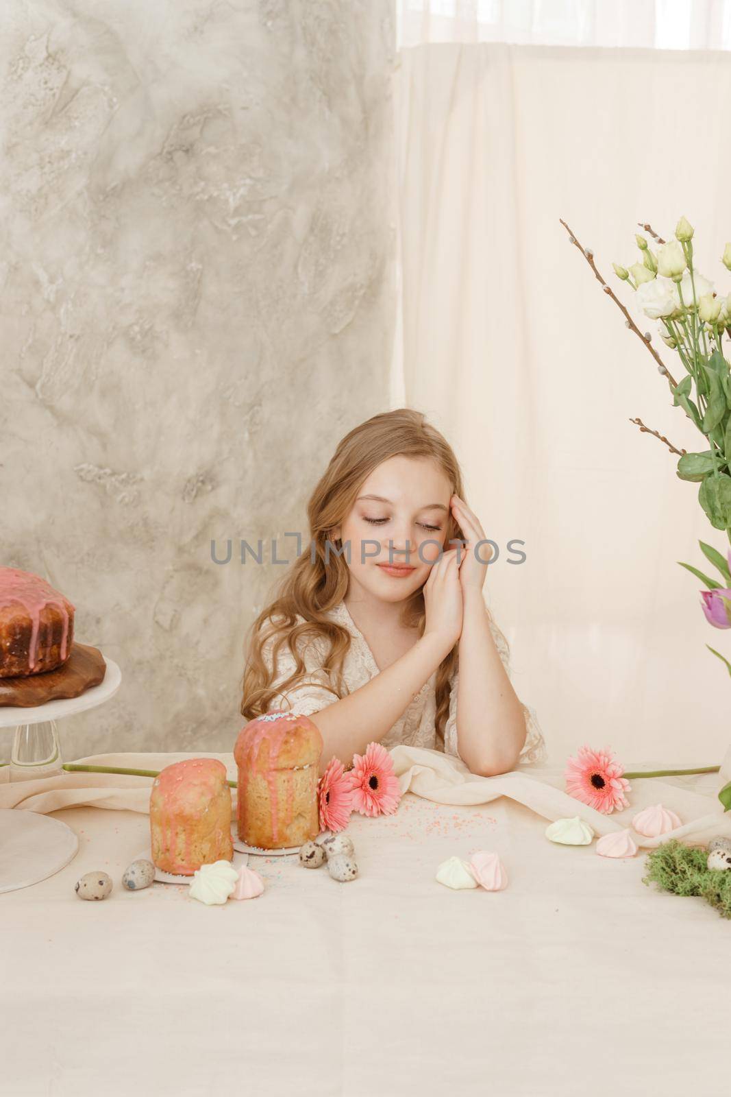 A girl with long hair in a light dress is sitting at the Easter table with cakes, spring flowers and quail eggs. Happy Easter celebration