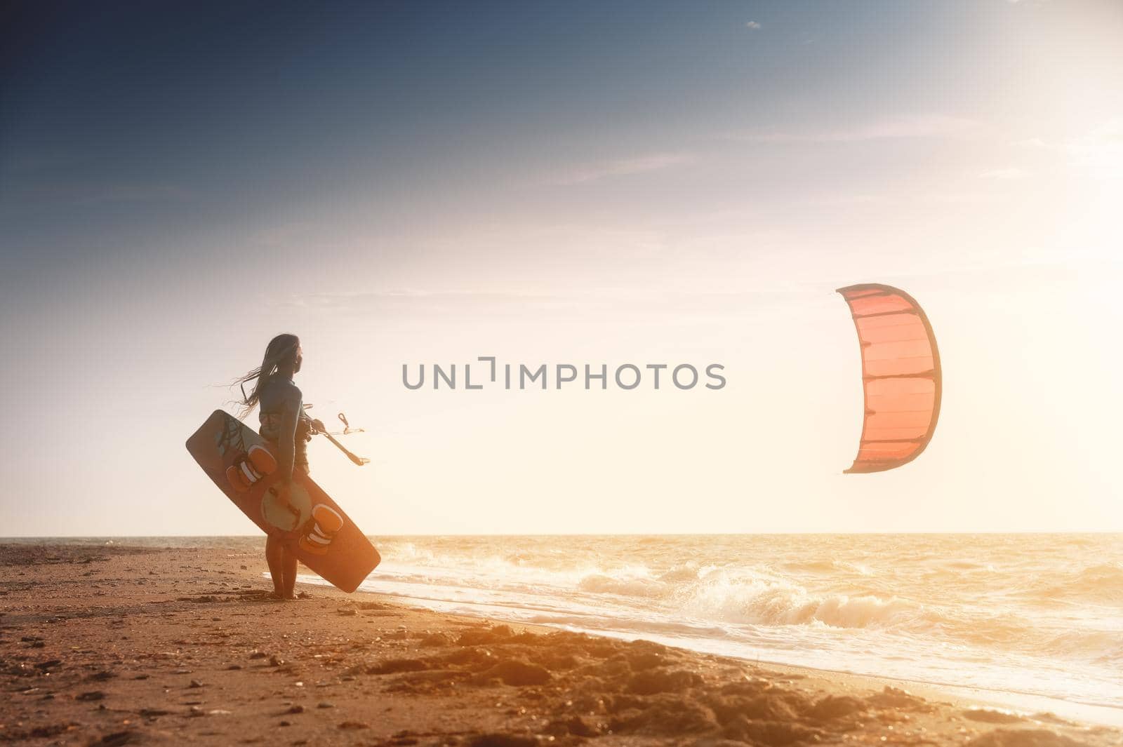 Kiteboarding. Kitesurfing athlete woman at sunset stands on the sandy shore holding her kite in the air and looks at the sea with waves and sunset by yanik88