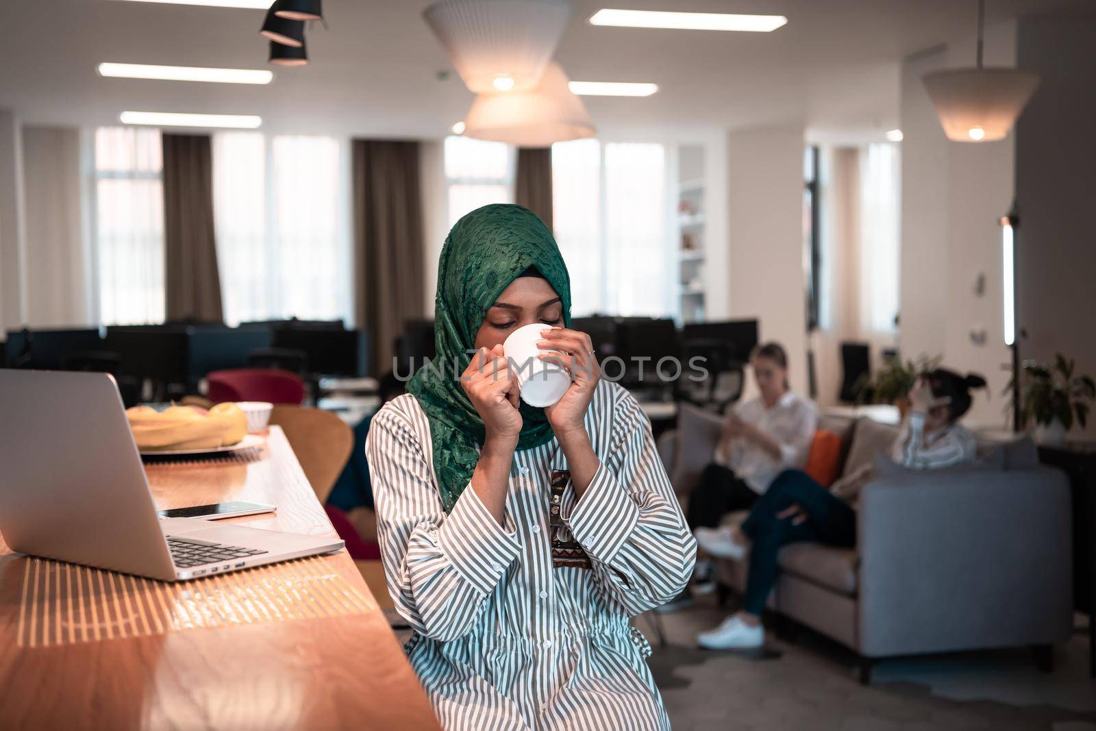 African Muslim businesswoman wearing a green hijab drinking tea while working on laptop computer in relaxation area at modern open plan startup office. High-quality photo