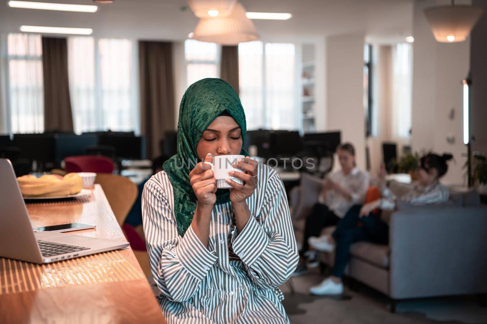 African Muslim businesswoman wearing a green hijab drinking tea while working on laptop computer in relaxation area at modern open plan startup office. High-quality photo