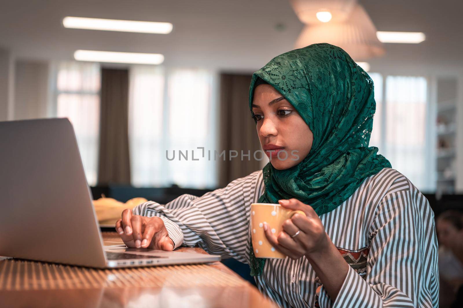 African Muslim businesswoman wearing a green hijab drinking tea while working on laptop computer in relaxation area at modern open plan startup office. High-quality photo