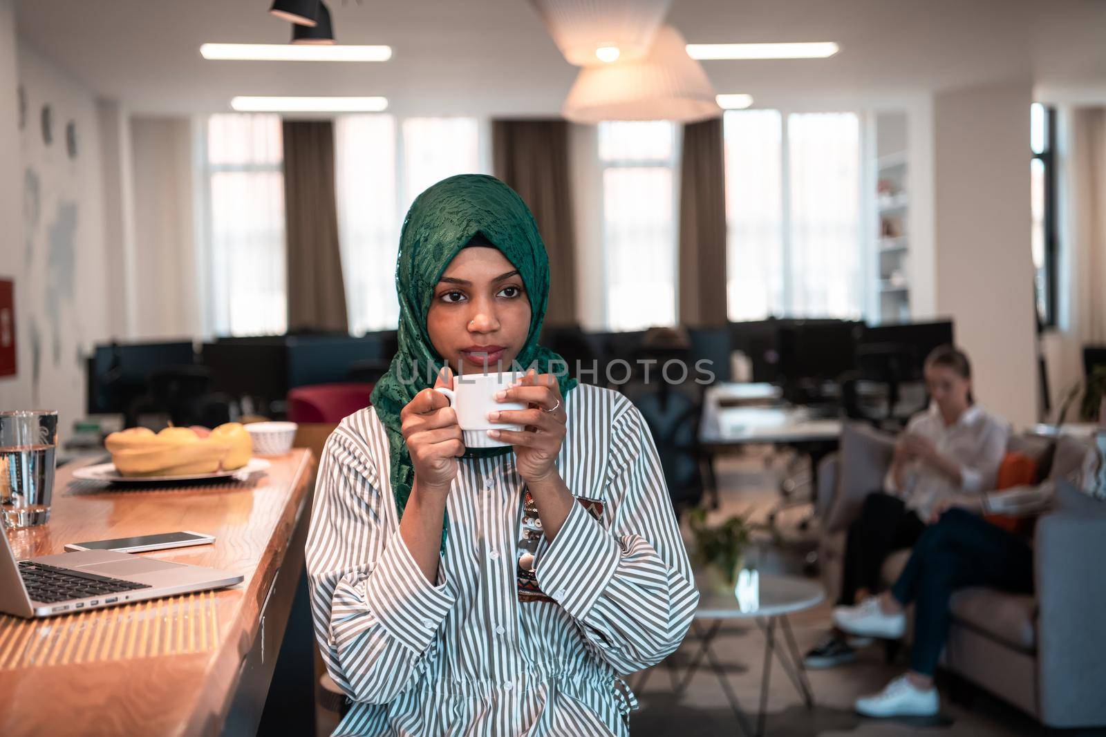 African muslim business woman wearing a green hijab drinking tea while working on laptop computer in relaxation area at modern open plan startup office. by dotshock