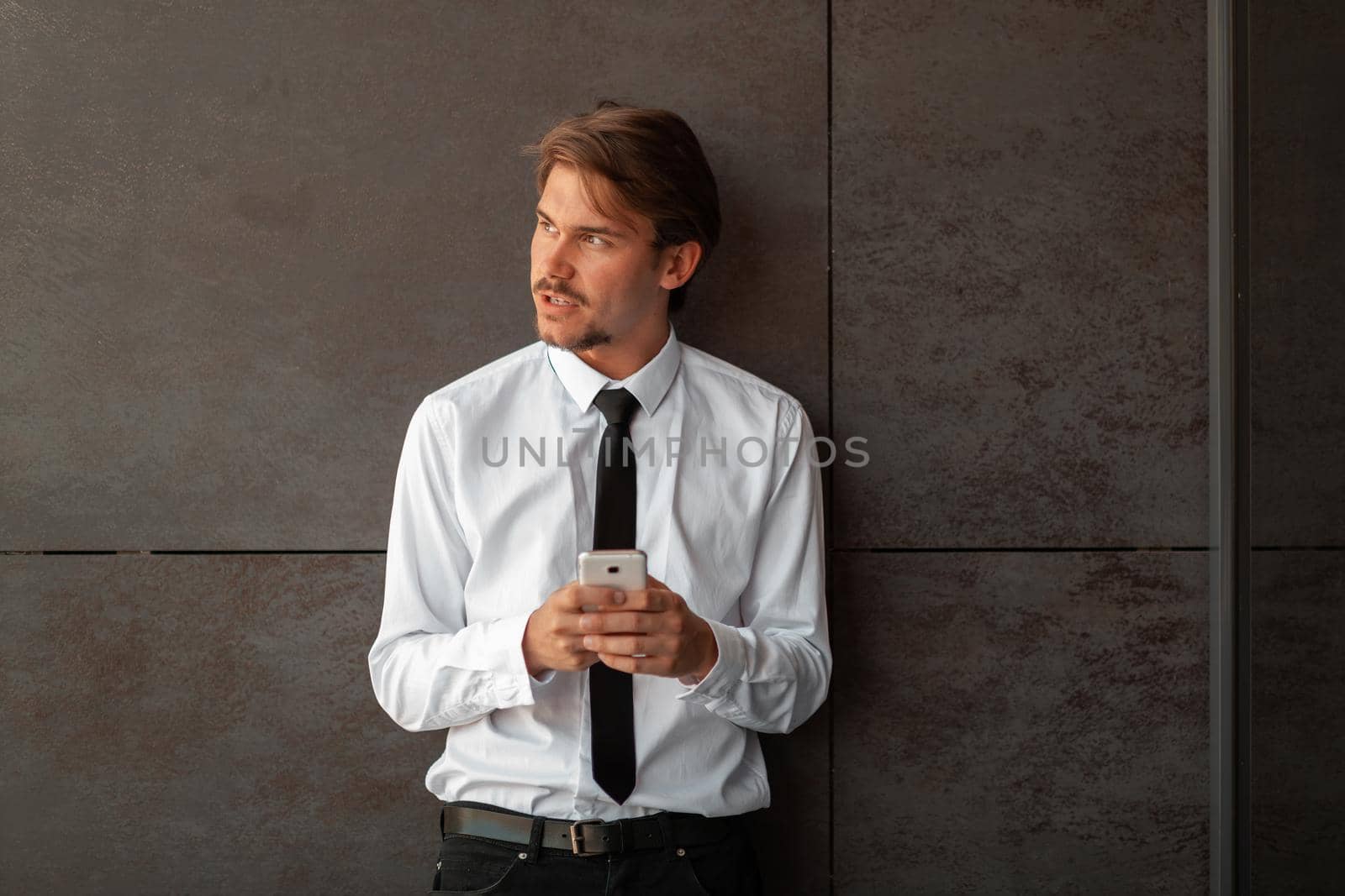 Startup businessman in a white shirt with a black tie using smartphone while standing in front of gray wall during break from work outside by dotshock