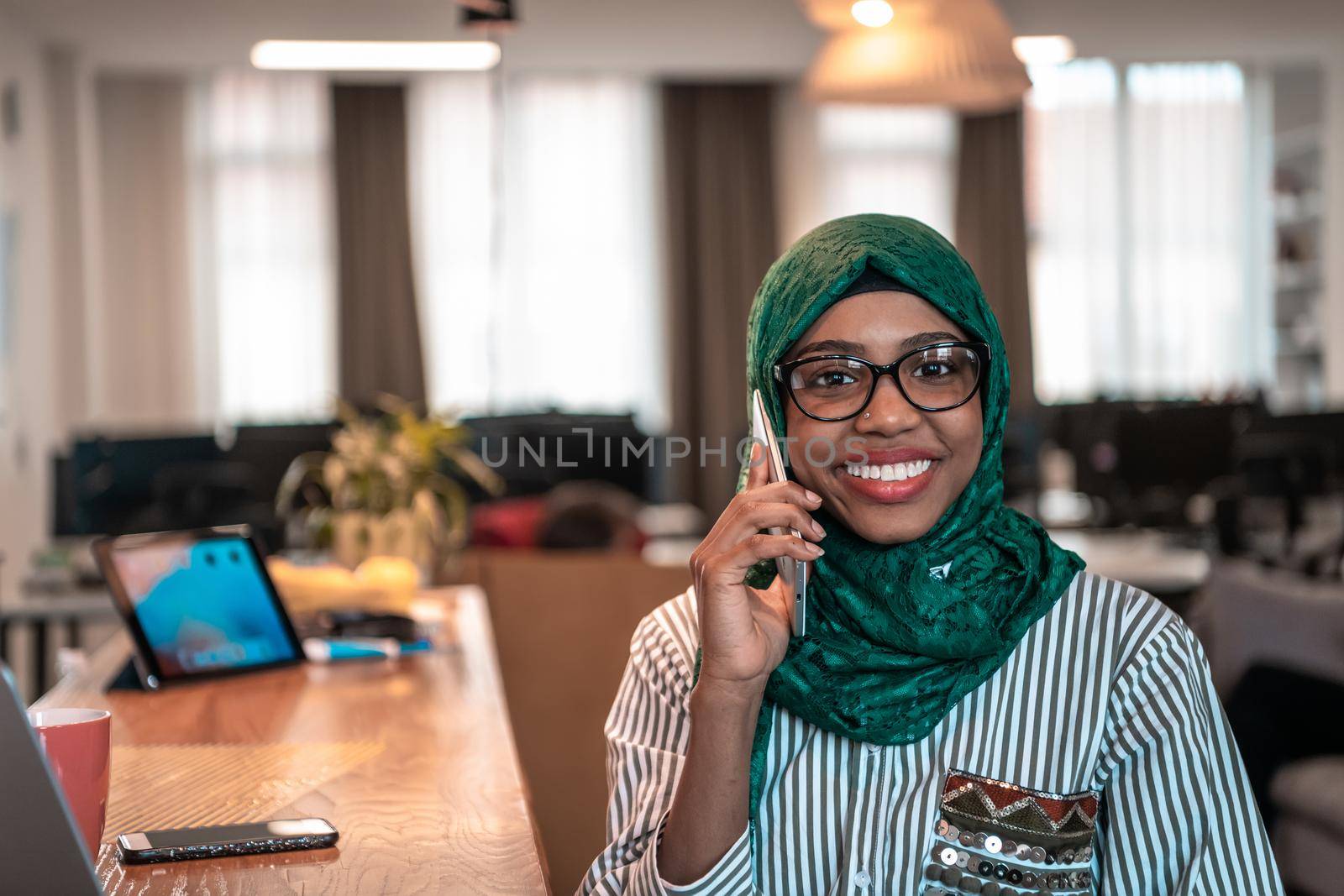 Smiling young woman talk on smartphone work on laptop look in distance thinking. Happy African American customer client use modern gadgets cellphone and computer at home office. by dotshock