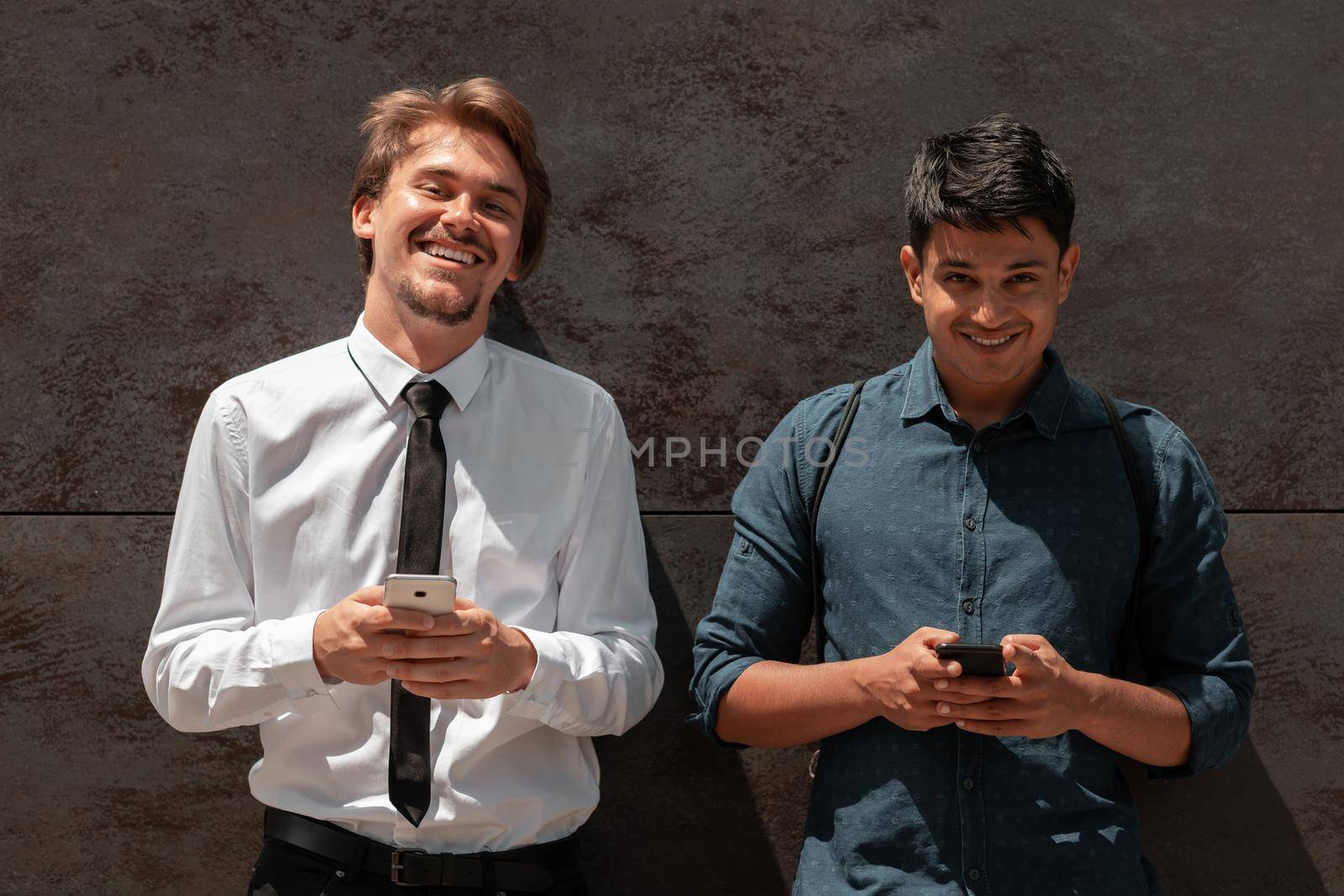 Casual multiethnic startup business men one of them is Indian using smartphone during break from work in front of grey wall outside by dotshock