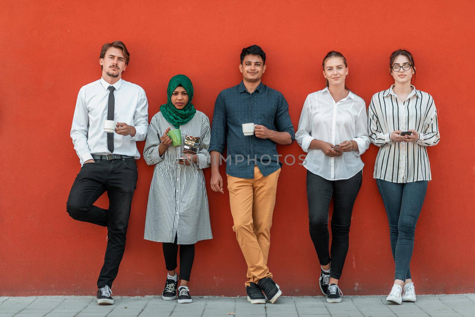 Multiethnic group of casual businesspeople using smartphone during a coffee break from work in front of the red wall outside. by dotshock