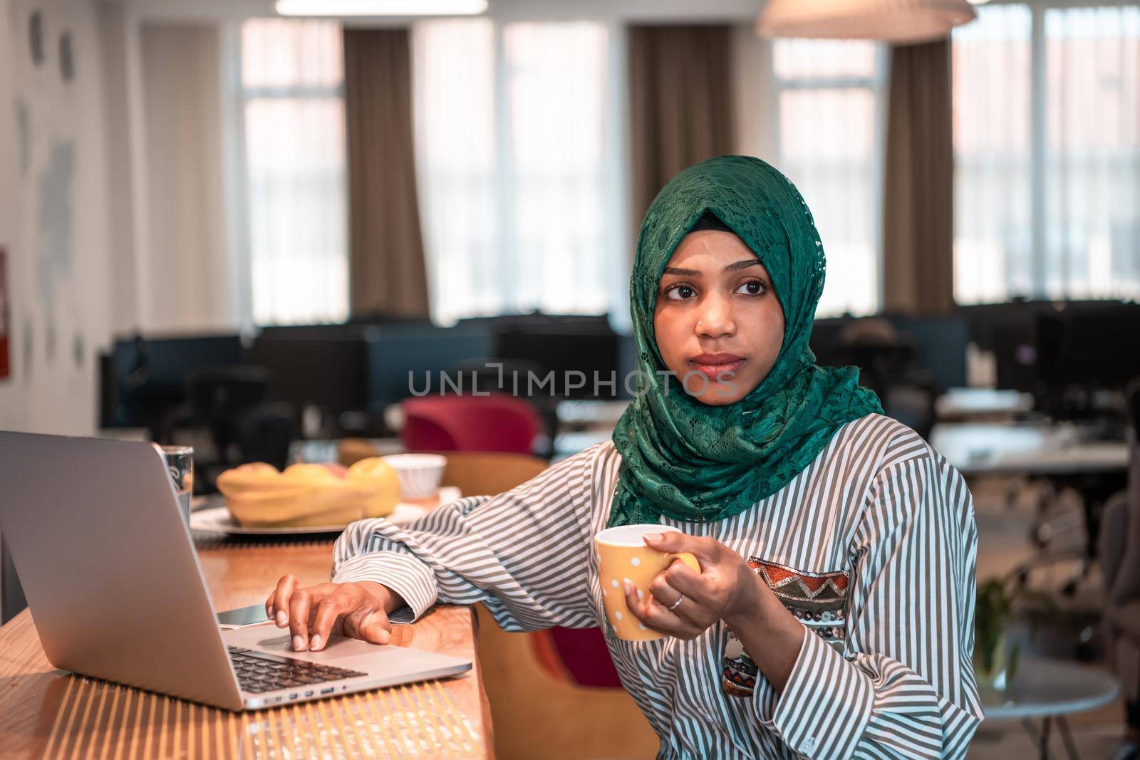 African Muslim businesswoman wearing a green hijab drinking tea while working on laptop computer in relaxation area at modern open plan startup office. High-quality photo