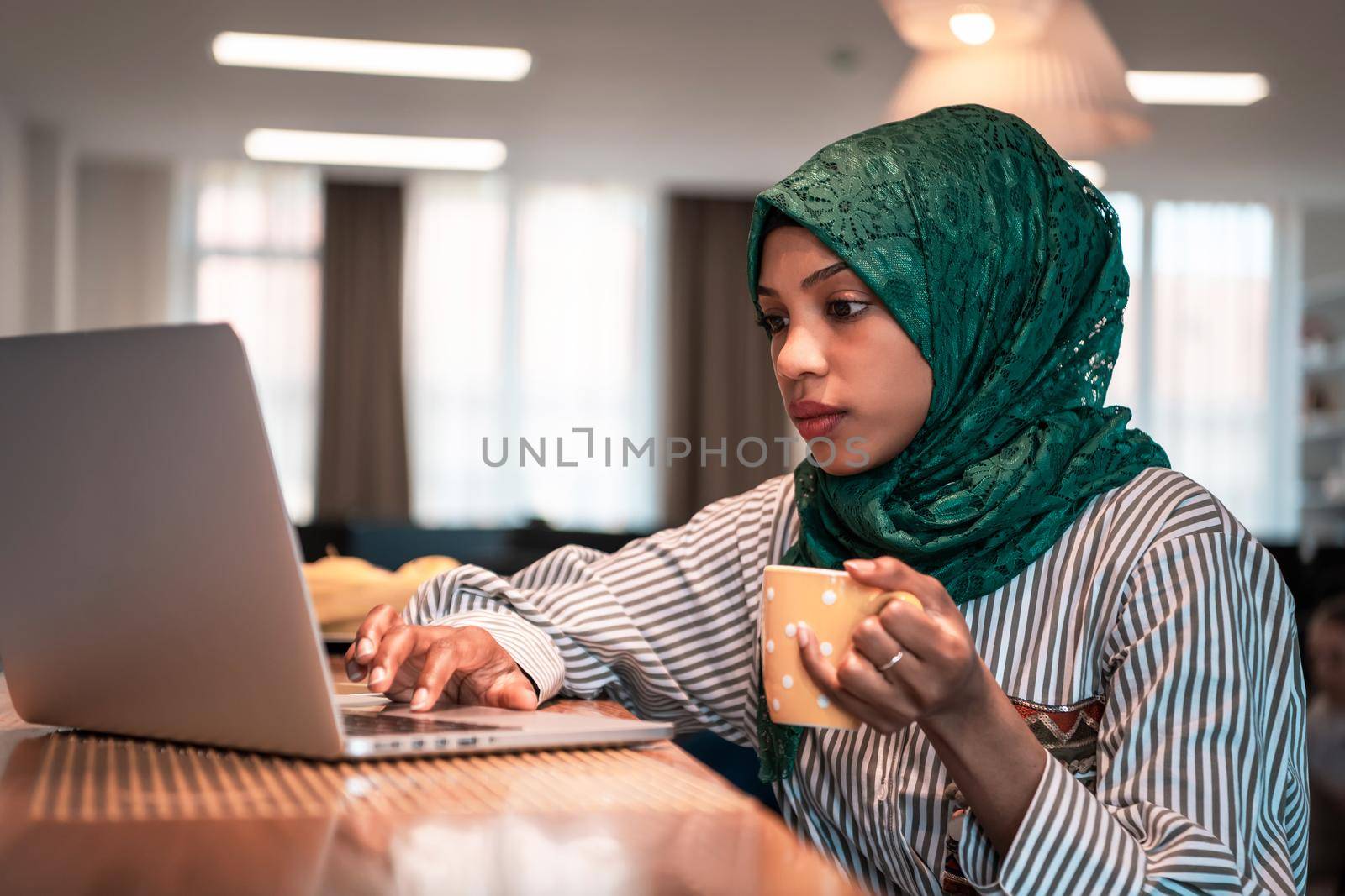 African Muslim businesswoman wearing a green hijab drinking tea while working on laptop computer in relaxation area at modern open plan startup office. High-quality photo