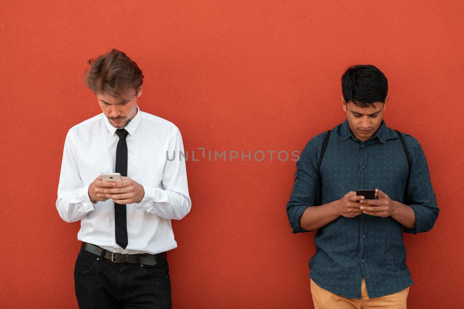 Casual multiethnic startup business men one of them is Indian using smartphone during a break from work in front of the red wall outside. High-quality photo