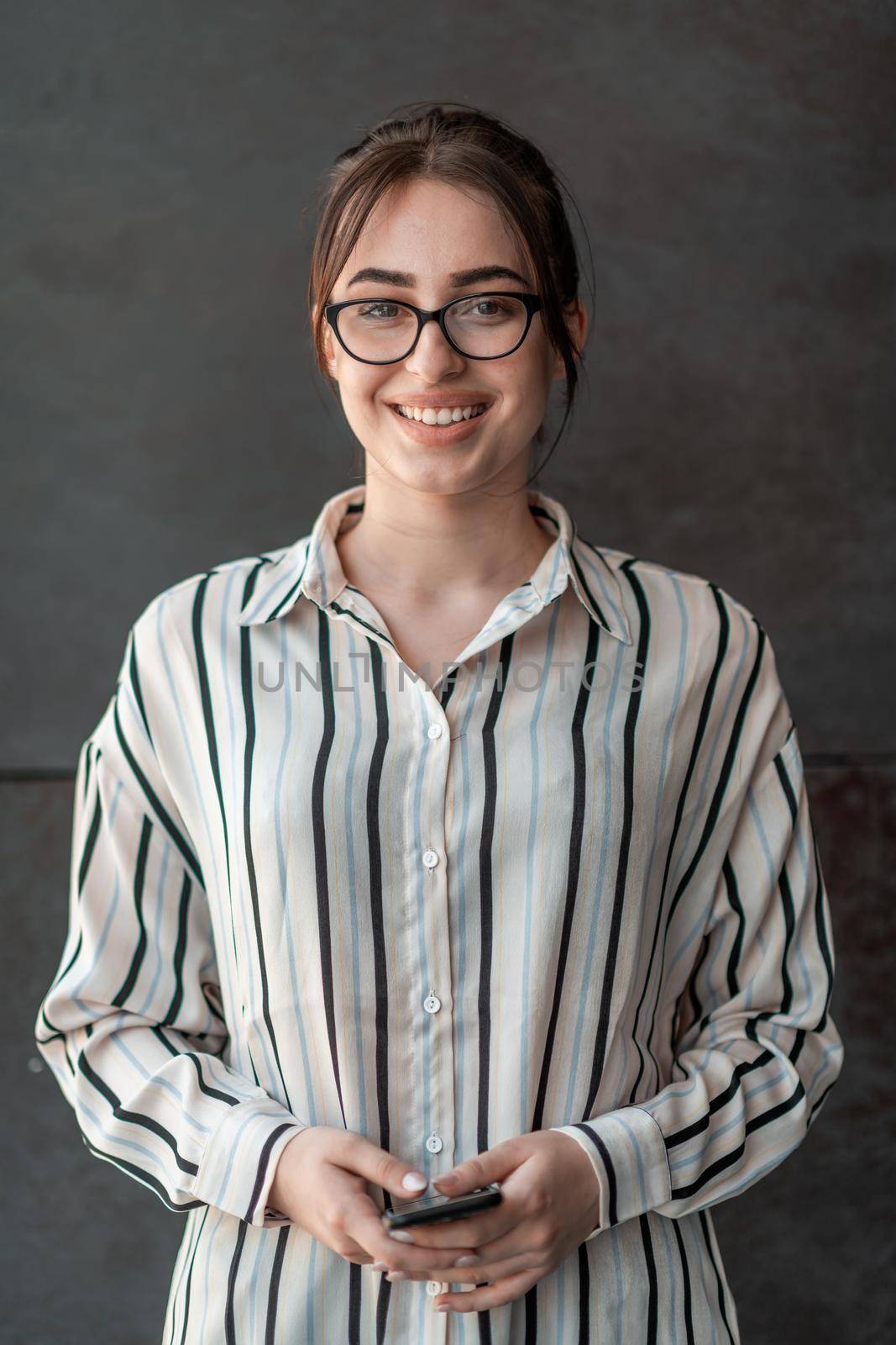 Startup businesswoman in shirt with a glasses using smartphone while standing in front of gray wall during break from work outside by dotshock