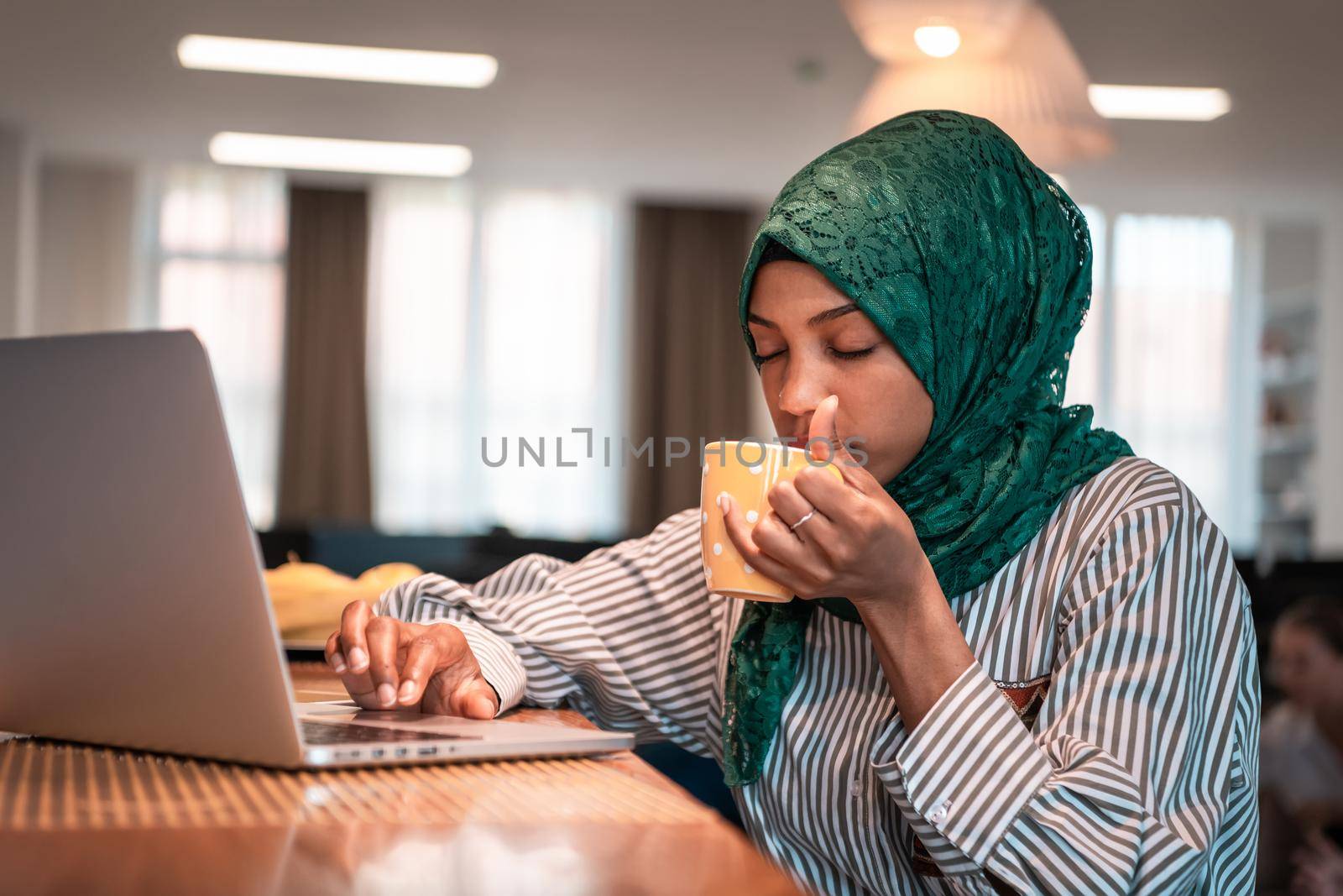 African Muslim businesswoman wearing a green hijab drinking tea while working on laptop computer in relaxation area at modern open plan startup office. High-quality photo