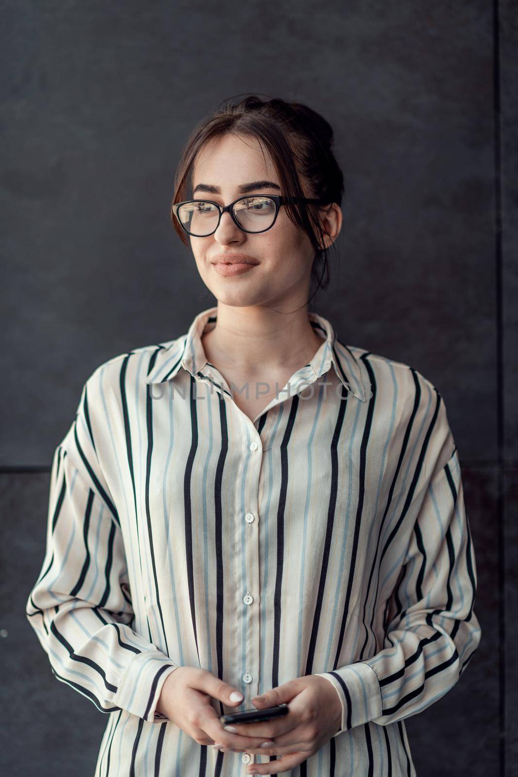 Startup businesswoman in shirt with a glasses using smartphone while standing in front of gray wall during a break from work outside. High-quality photo
