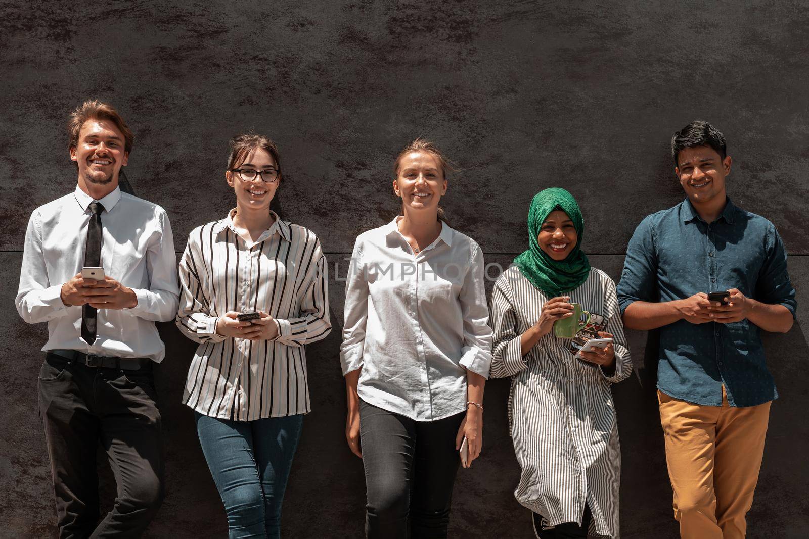 Multiethnic group of casual businesspeople using a smartphone during a coffee break from work in front of the black wall outside. High-quality photo