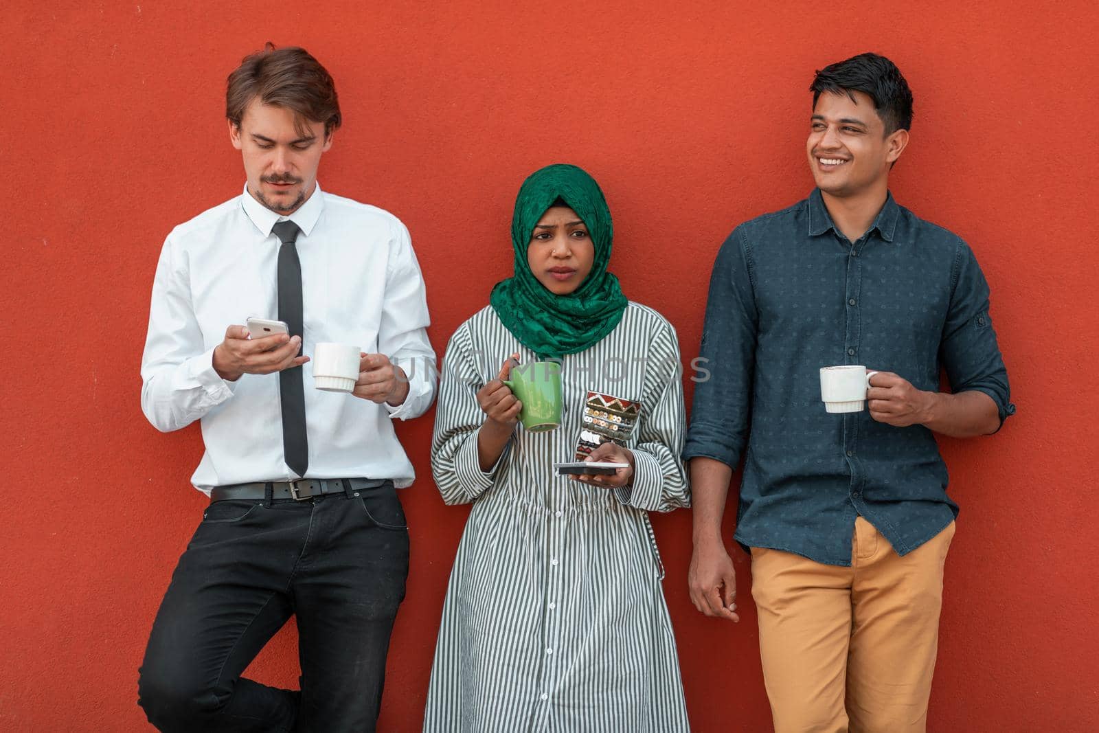 Multiethnic group of casual businesspeople using smartphones during a coffee break from work in front of the red wall outside. High-quality photo