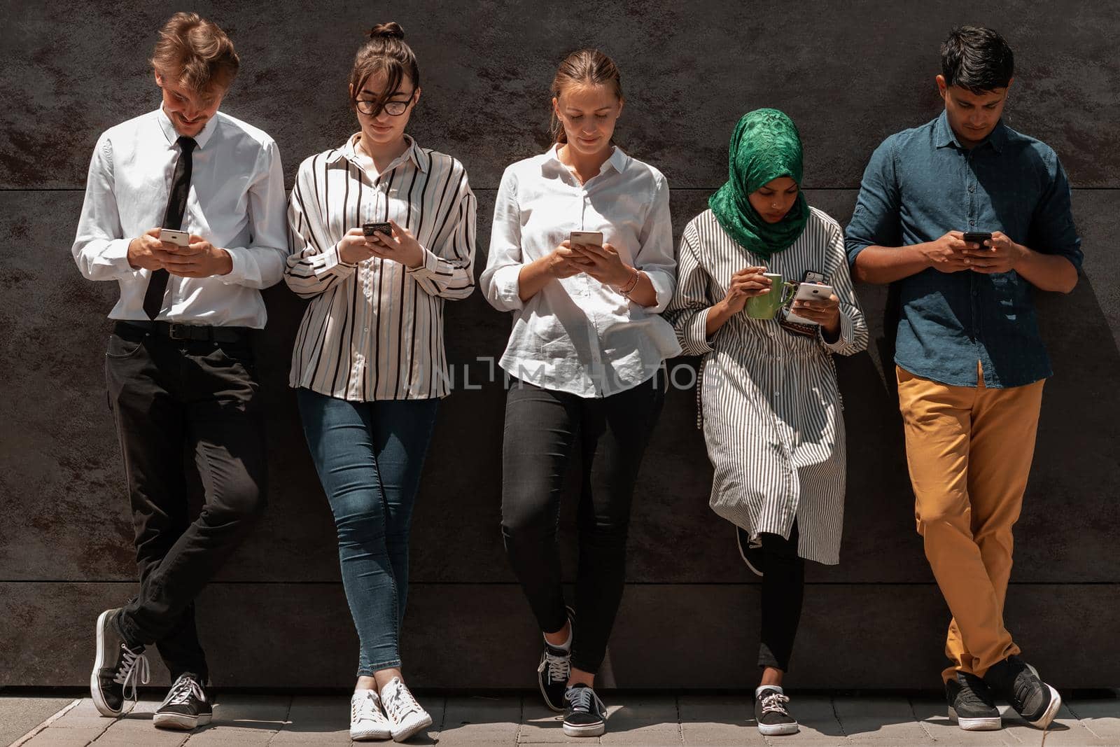 Multiethnic group of casual businesspeople using a smartphone during a coffee break from work in front of the black wall outside. High-quality photo