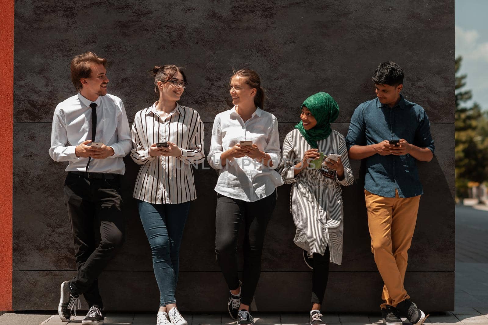 Multiethnic group of casual businesspeople using a smartphone during a coffee break from work in front of the black wall outside. High-quality photo