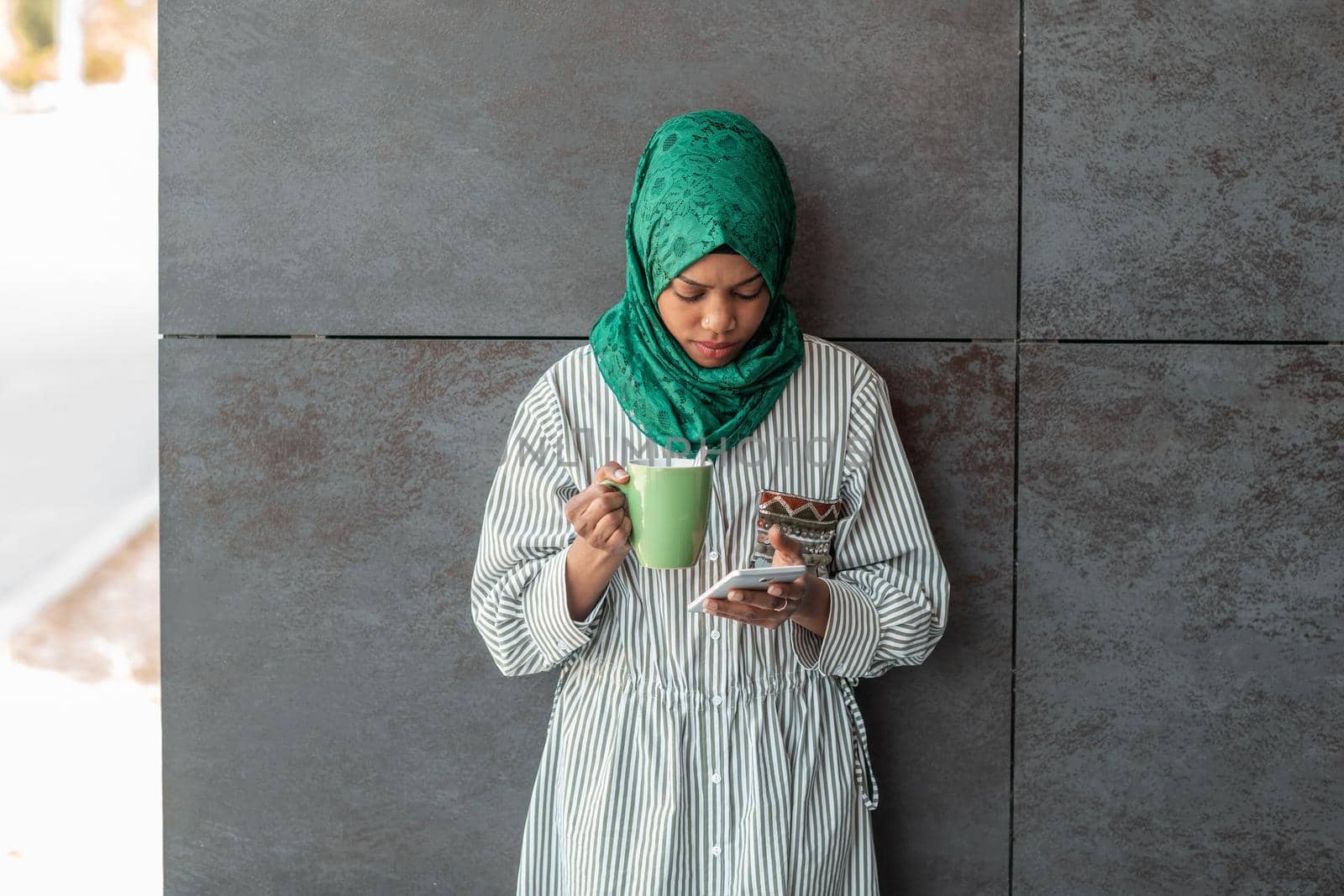 African Muslim businesswoman with green hijab using a smartphone during a coffee break from work outside. High-quality photo