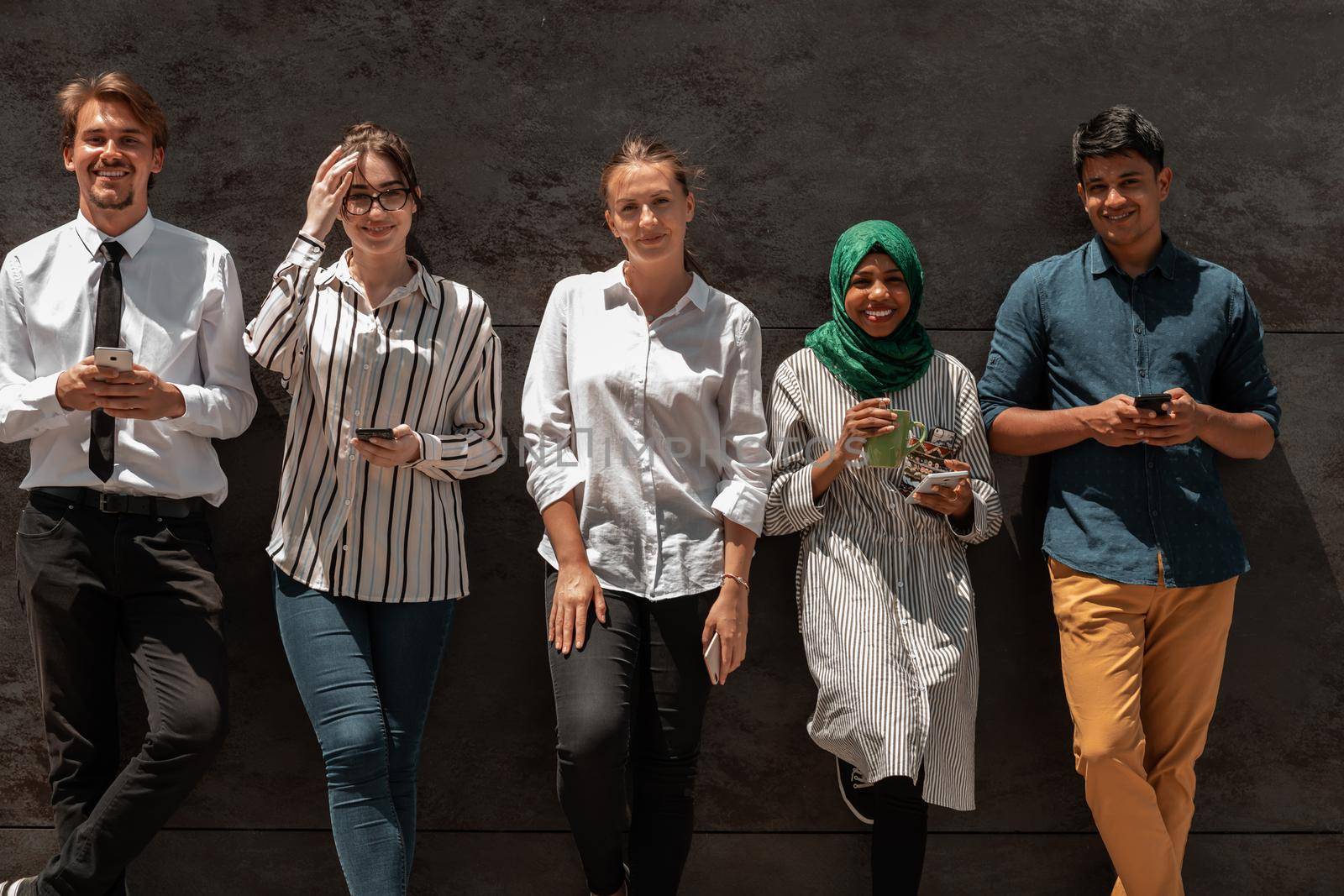 Multiethnic group of casual businesspeople using a smartphone during a coffee break from work in front of the black wall outside. High-quality photo