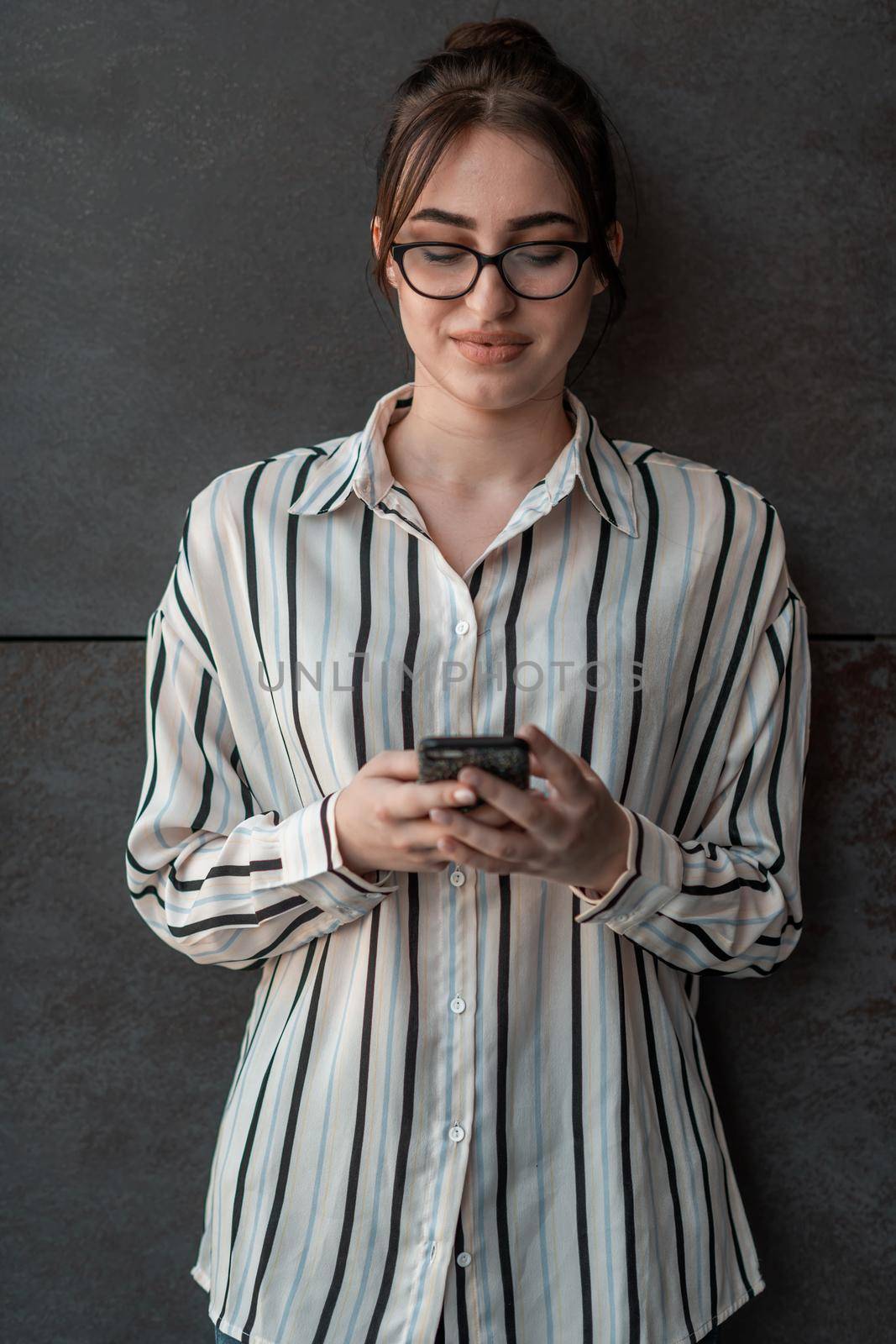 Startup businesswoman in shirt with a glasses using smartphone while standing in front of gray wall during a break from work outside. High-quality photo