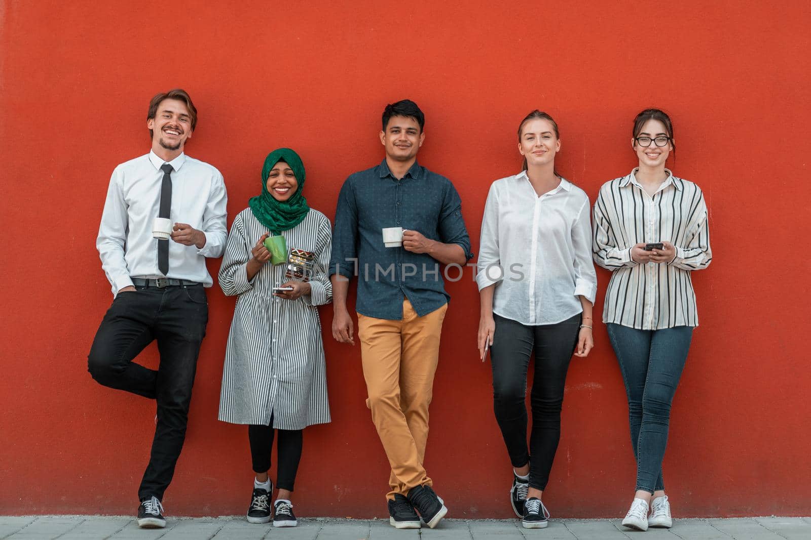 Multiethnic group of casual businesspeople using smartphone during a coffee break from work in front of the red wall outside. by dotshock