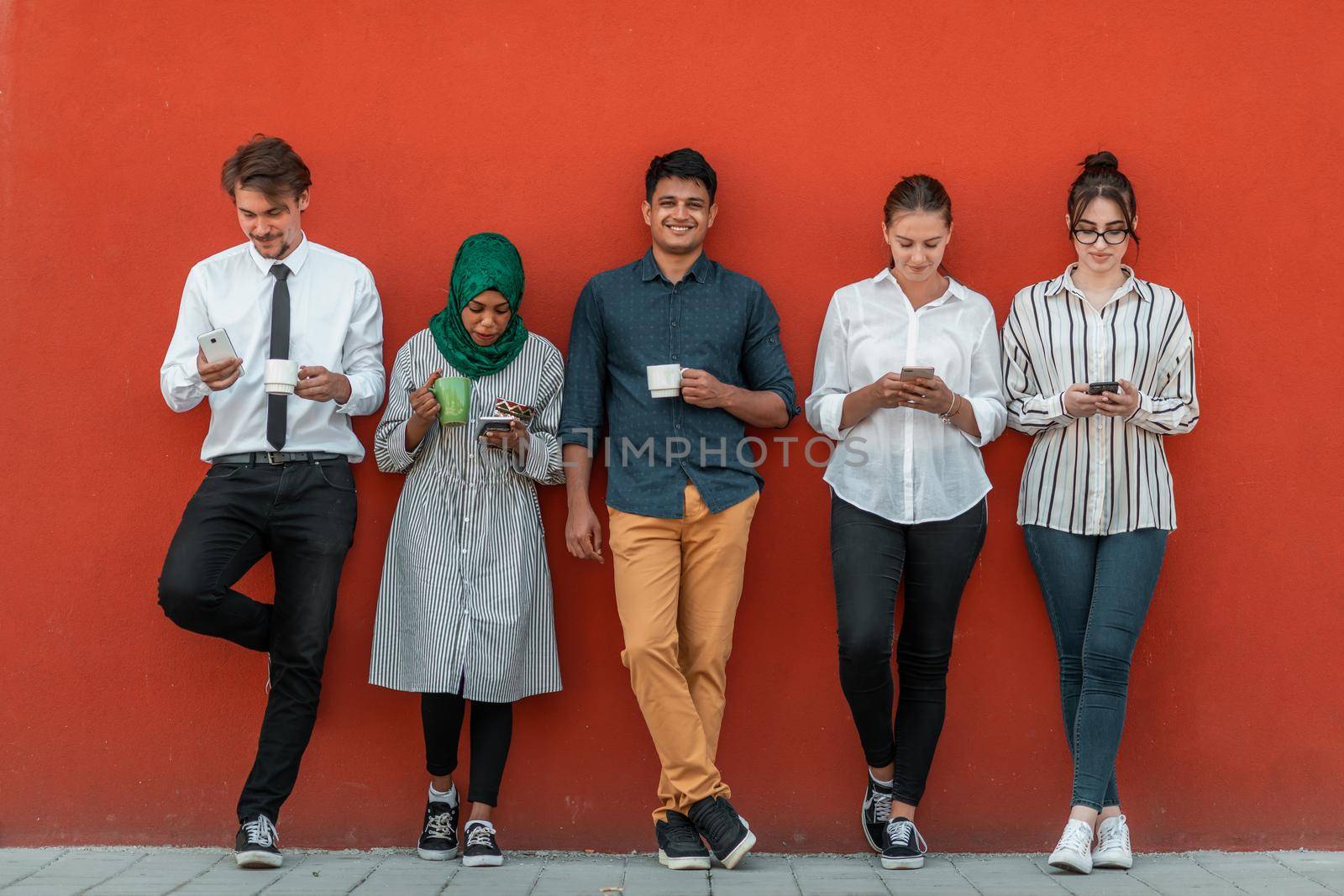 Multiethnic group of casual businesspeople using smartphones during a coffee break from work in front of the red wall outside. High-quality photo