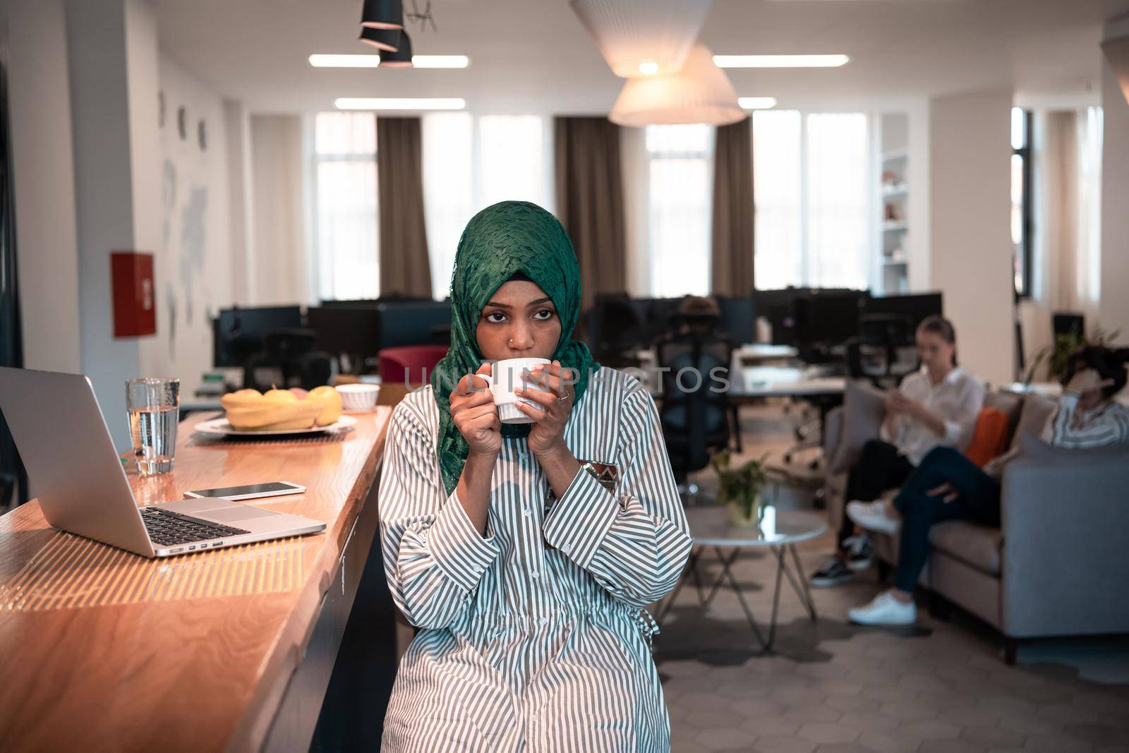 African Muslim businesswoman wearing a green hijab drinking tea while working on laptop computer in relaxation area at modern open plan startup office. High-quality photo