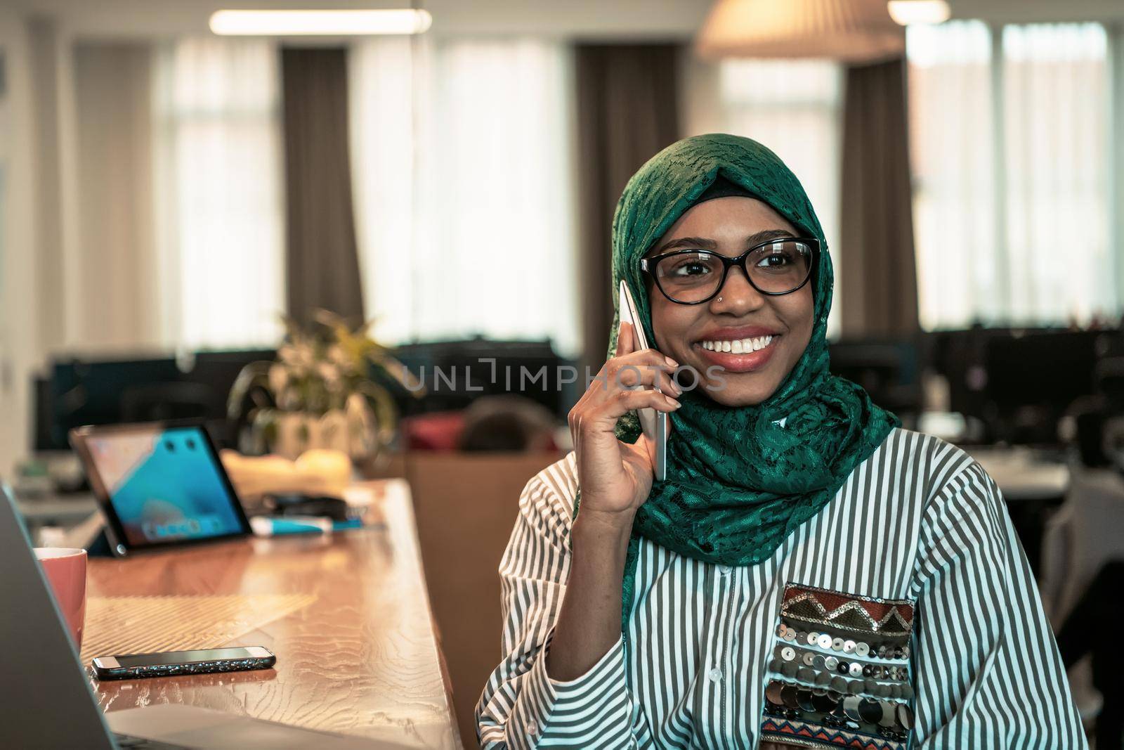 Smiling young woman talk on smartphone work on laptop look in distance thinking. Happy African American customer client using modern gadgets cellphone and computer at the home office. High-quality photo