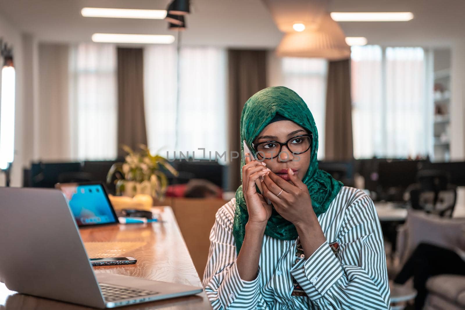 Smiling young woman talk on smartphone work on laptop look in distance thinking. Happy African American customer client use modern gadgets cellphone and computer at home office. by dotshock