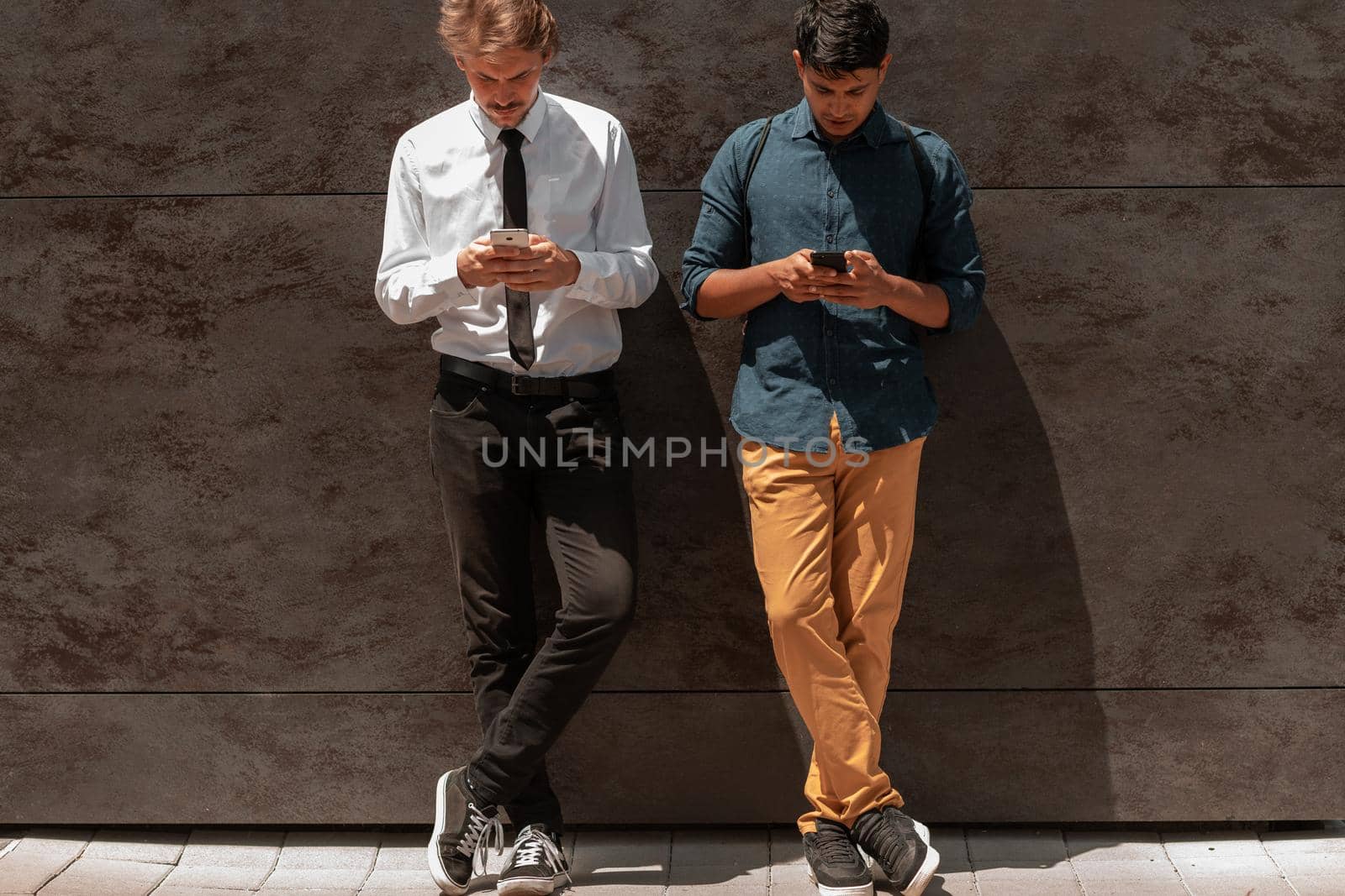 Casual multiethnic startup business men one of them is Indian using smartphone during break from work in front of grey wall outside by dotshock