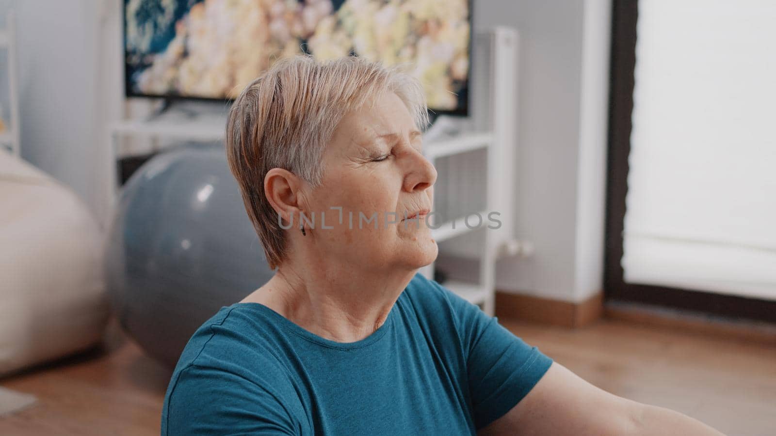 Close up of pensioner meditating on floor mat at home, having eyes closed in concentration. Senior woman doing relaxing meditation before training with physical exercise and gymnastics.