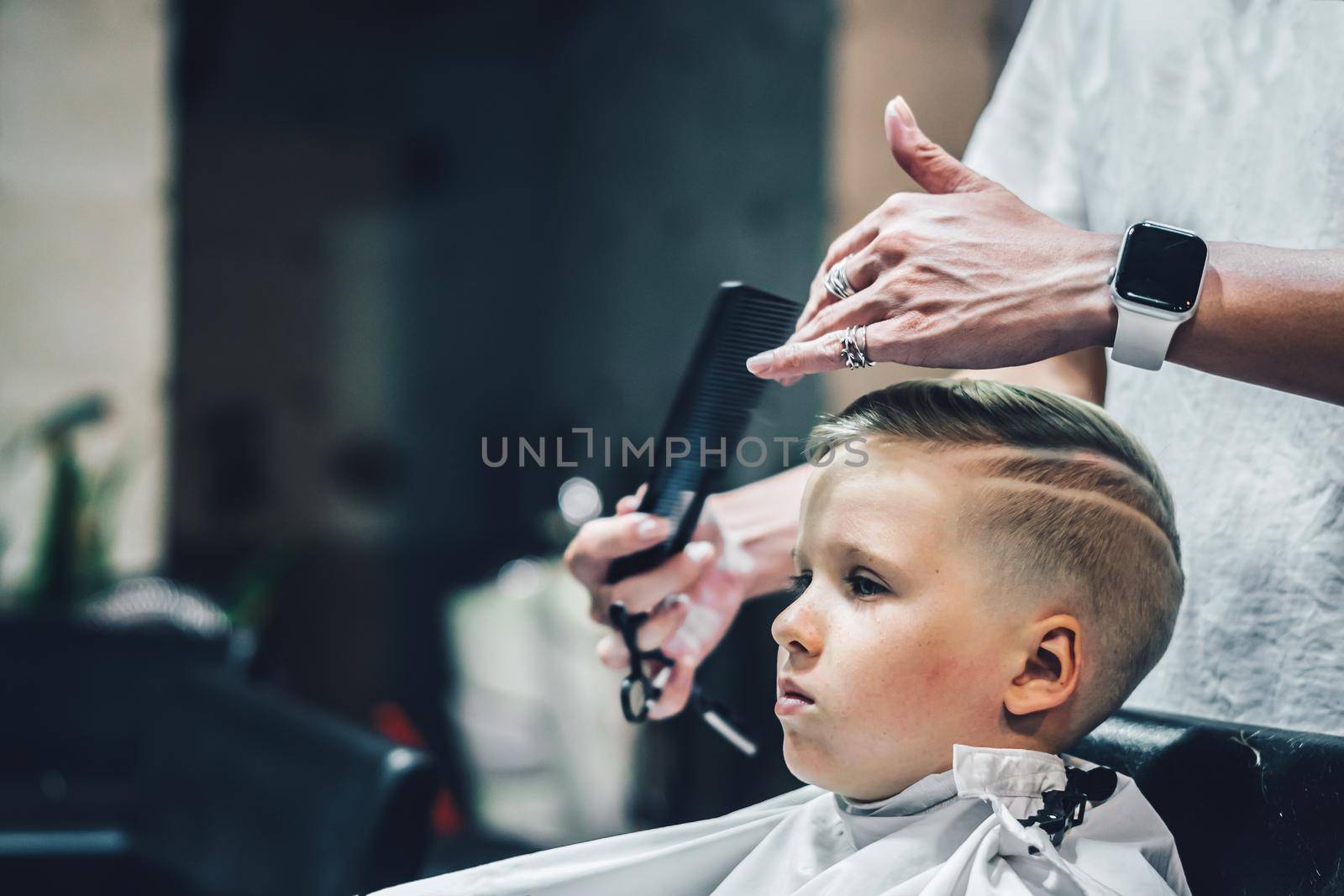 Lifestyle photo Barber shop blond boy customer. Hairdresser man work move hand comb tool scissors dryer clip white cape. Modern style kid short hair cut. Fashion business service for children.