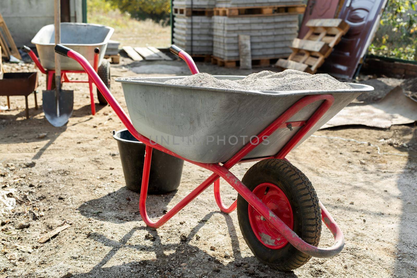 Construction wheelbarrow with cement and a shovel on the background of a stack of gray tiles by StudioPeace