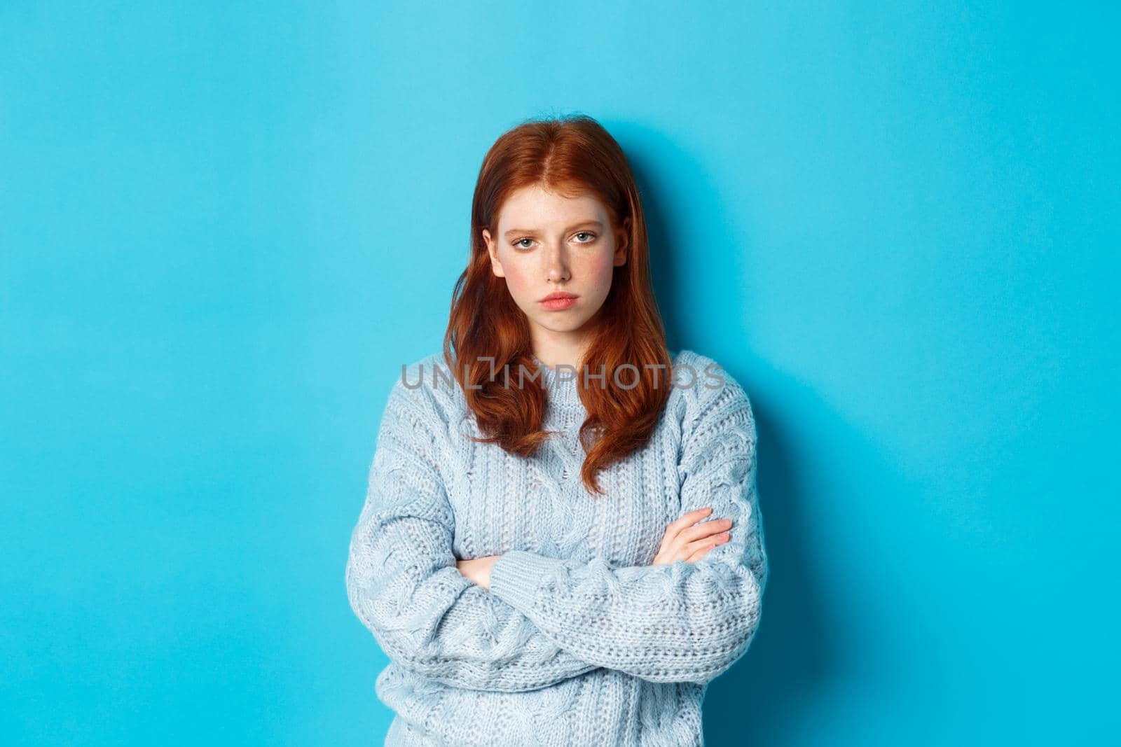 Annoyed and bothered redhead teeange girl cross arms on chest, staring at something lame and boring, standing against blue background.