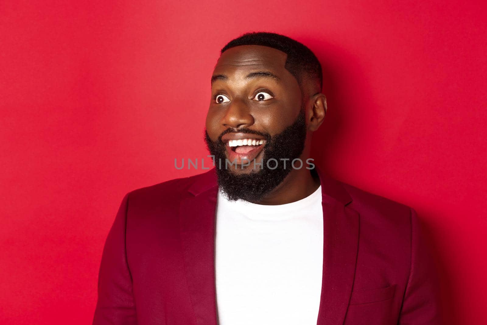 Close-up of excited african american guy looking left, smiling with amazement and joy, standing in blazer against red background by Benzoix