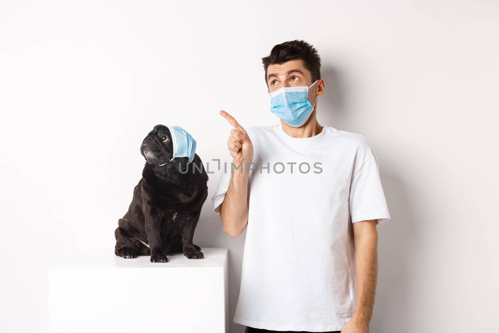 Covid-19, animals and quarantine concept. Young man and black dog wearing medical masks, pug and owner looking at upper left corner, white background by Benzoix