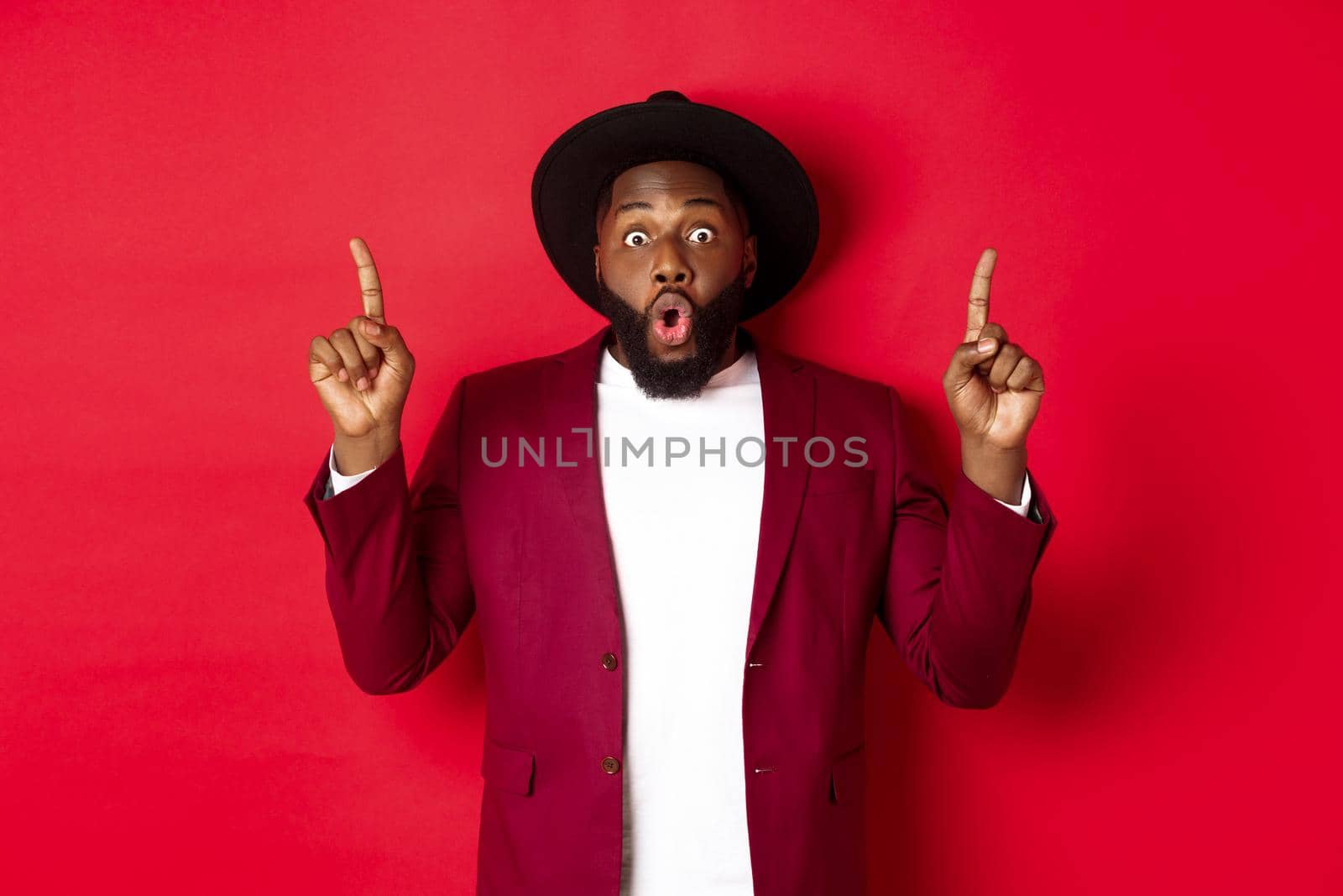 Winter holidays and shopping concept. Impressed african american man in party outfit pointing fingers up, checking out christmas promo, standing over red background.