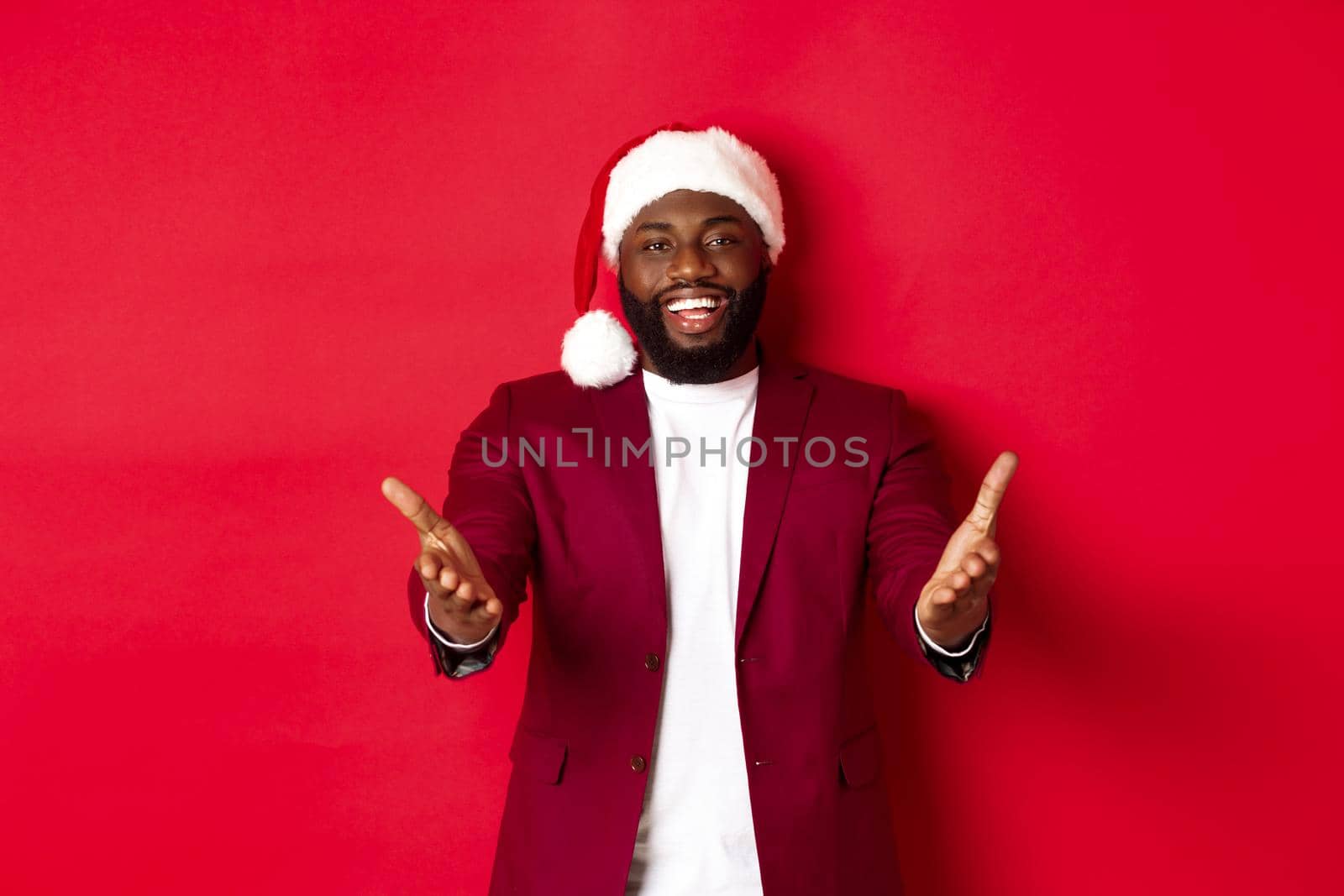 Christmas, party and holidays concept. Happy Black man in santa hat welcome you, pointing hands at camera with pleased smile, standing against red background.