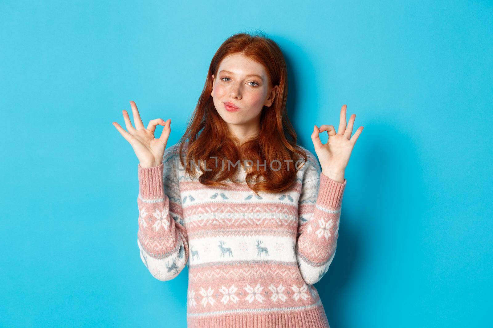 Satisfied and proud redhead girl nod in approval, showing okay sign, not bad or praise gesture, standing against blue background.