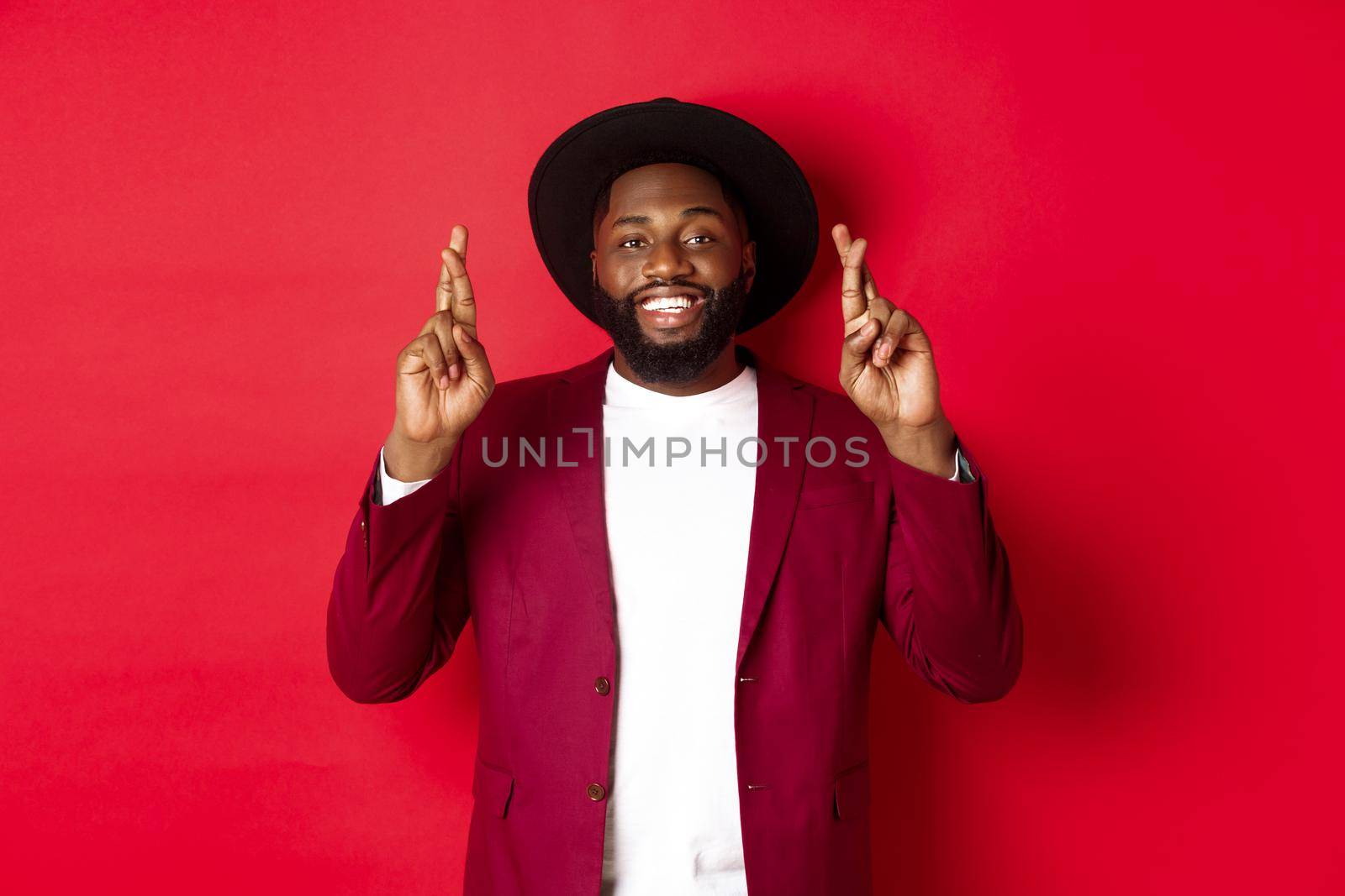 Hopeful african american man making wish, holding fingers crossed for good luck and smiling optimistic, standing against red party background.