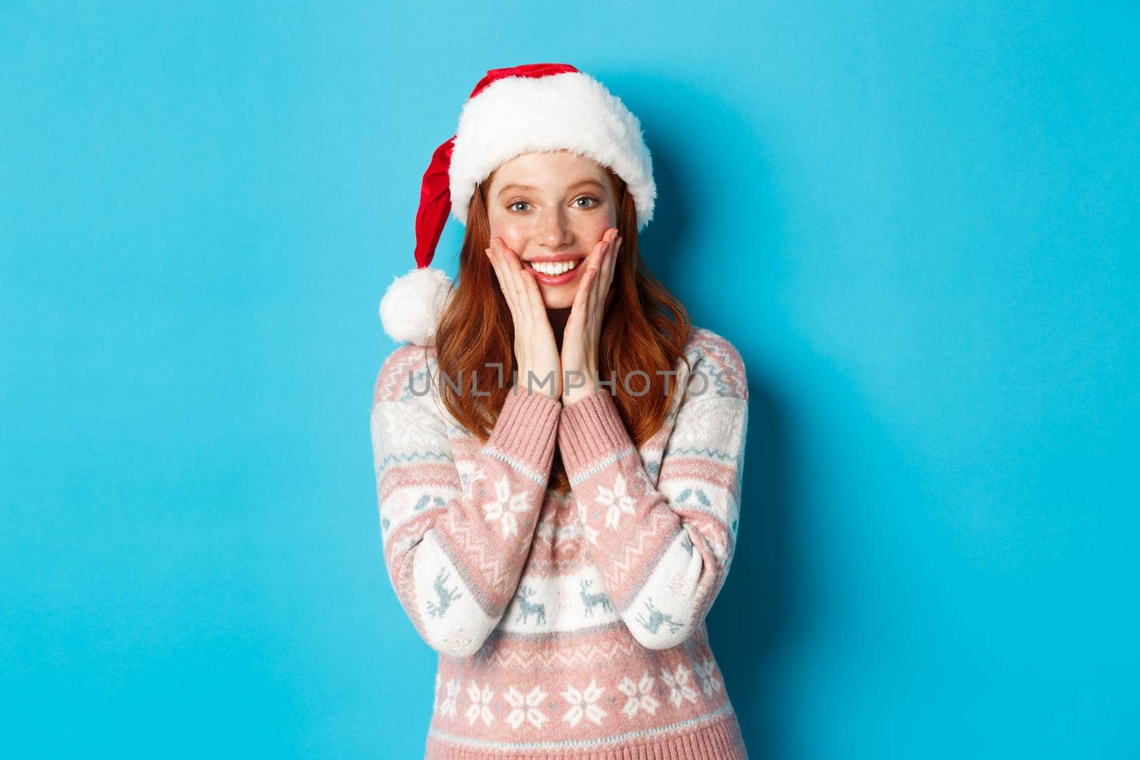 Winter and Christmas Eve concept. Happy redhead girl celebrating xmas, staring at camera amazed, smiling and touching cheeks, standing over blue background by Benzoix