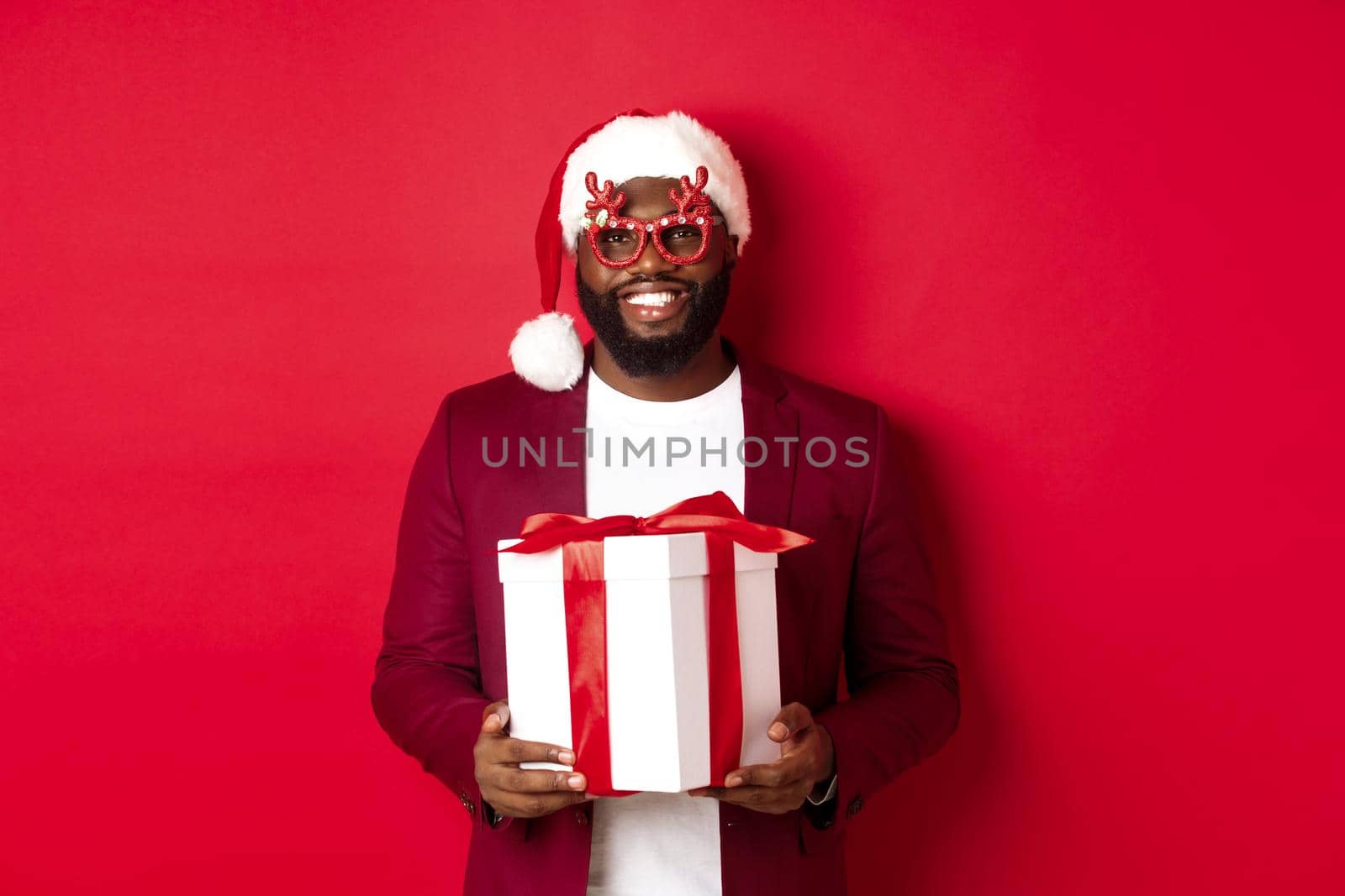Christmas. Handsome african american man in party glasses and santa hat holding new year gift, bring present in box and smiling, standing over red background.
