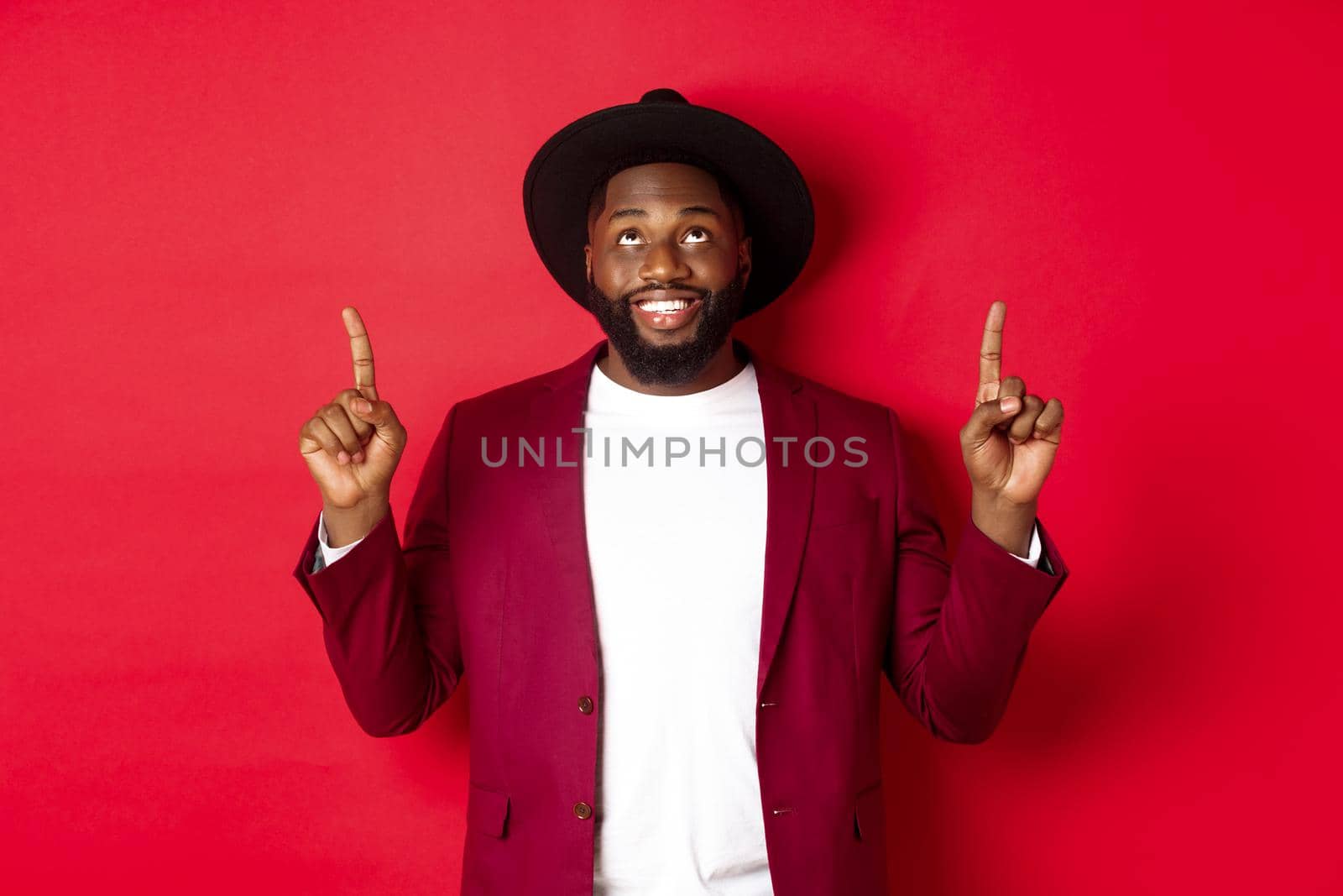 Winter holidays and shopping concept. Happy african american man wearing party clothing for new year, pointing and looking up with pleased smile, red background by Benzoix