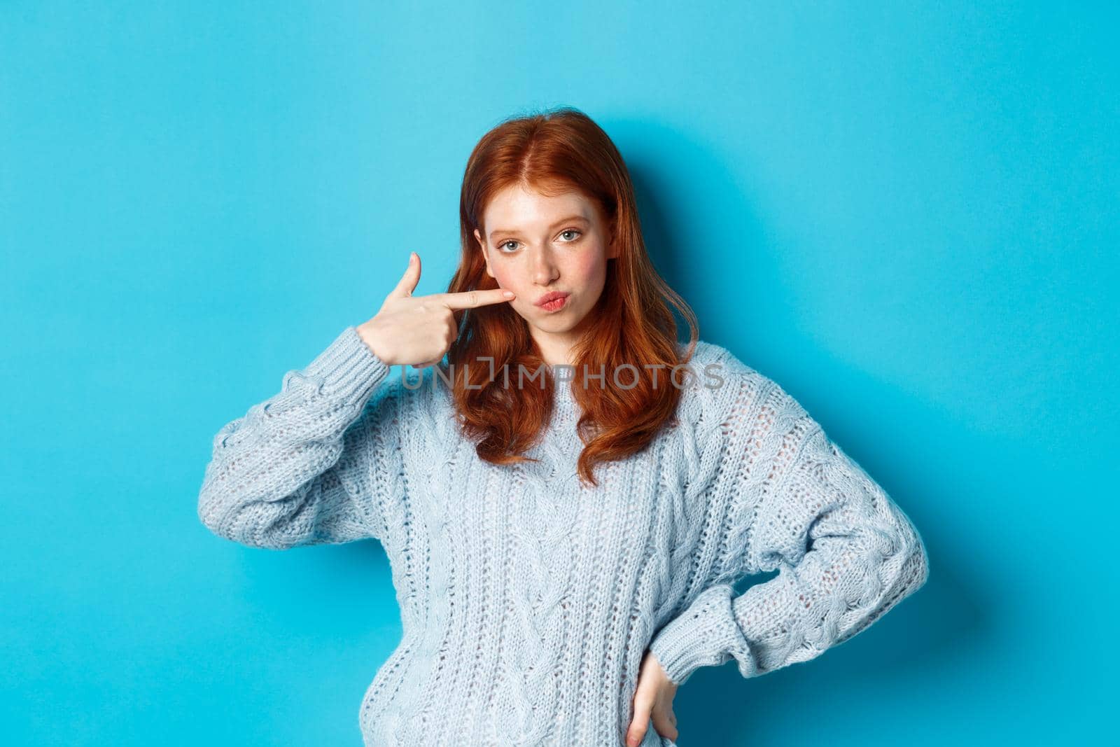 Cute redhead girl in sweater poking her cheek, staring at camera sassy, standing over blue background.