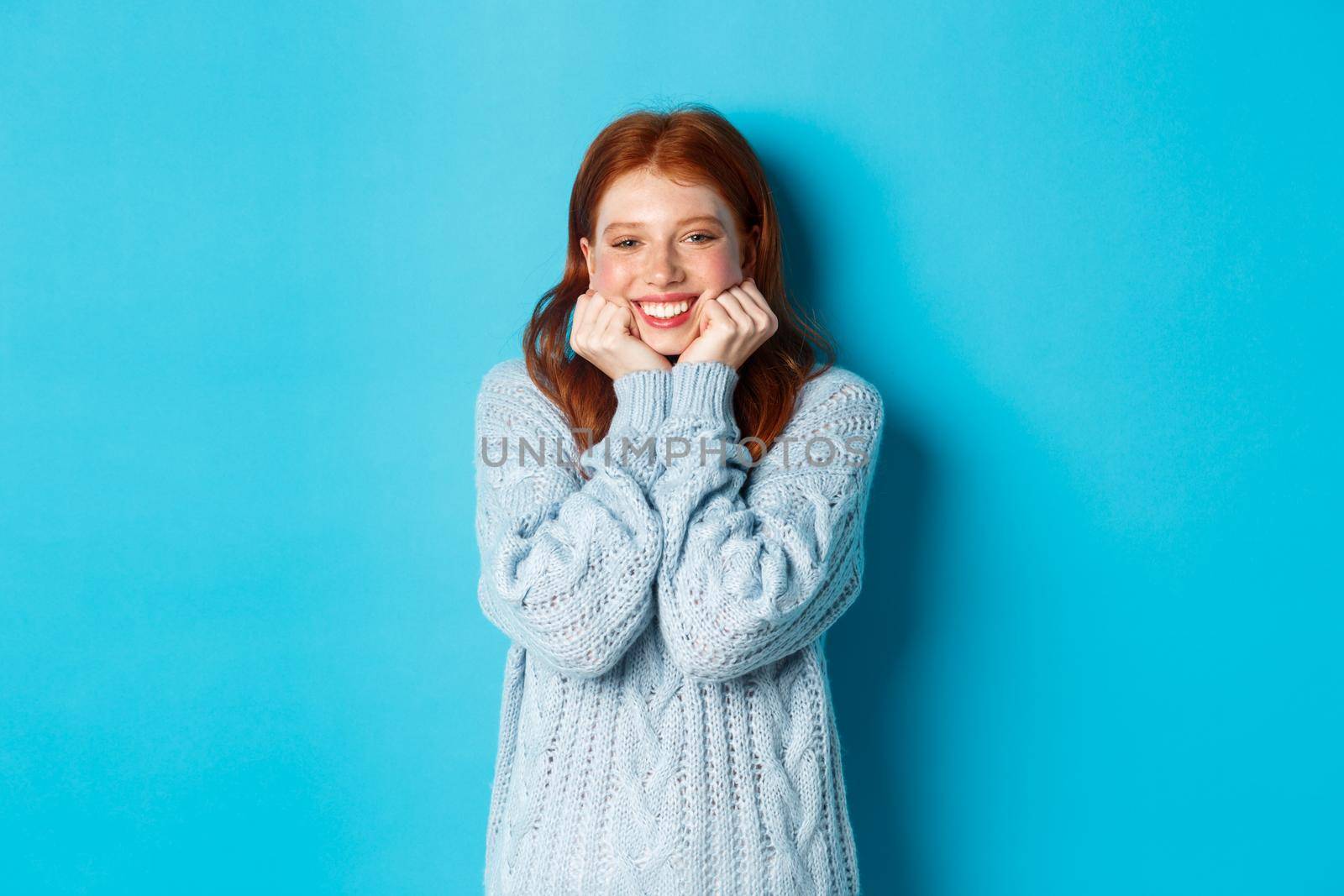 Lovely redhead teenage girl looking daydreaming, staring with admirationa sympathy at camera, standing against blue background.