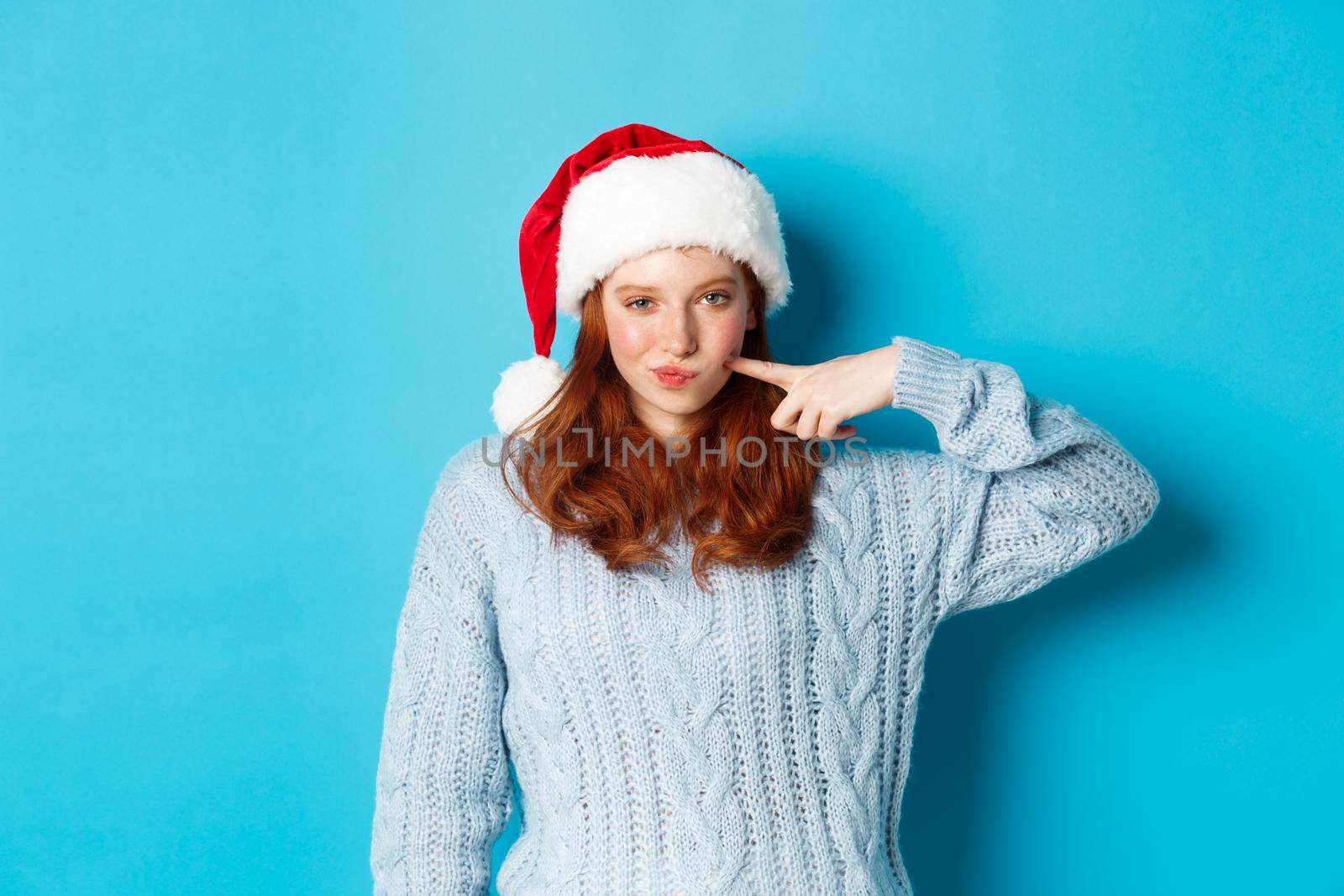 Winter holidays and Christmas Eve concept. Sassy teenage girl with red hair, wearing santa hat, enjoying New Year, poking her cheek and looking confident at camera.